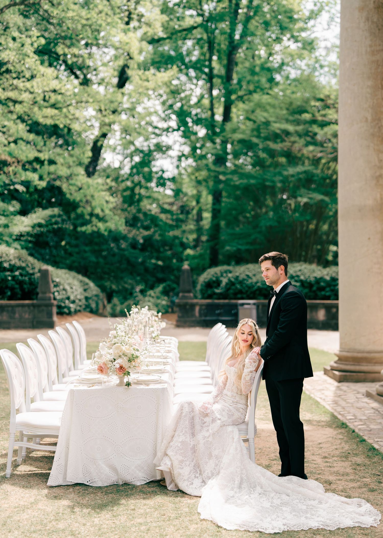 Elegant bride in lace gown seated at outdoor wedding reception table with groom in tuxedo amid lush garden setting.