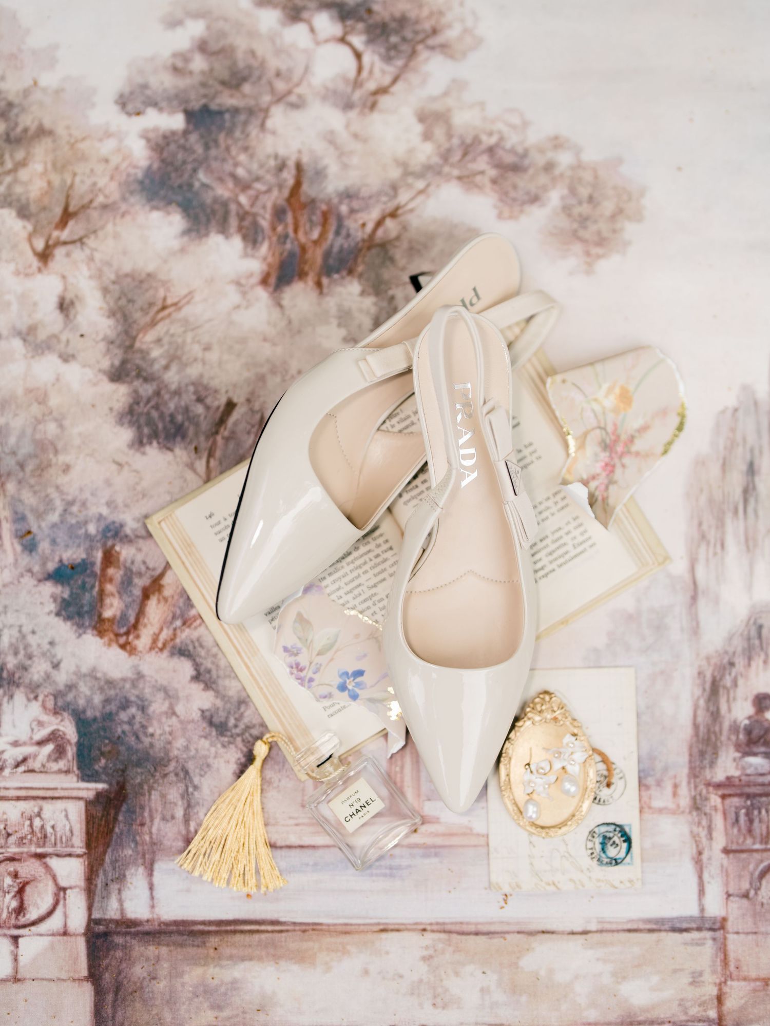 Elegant cream Dior bridal flats displayed with perfume, vintage brooch, and tassel on floral wallpaper backdrop.