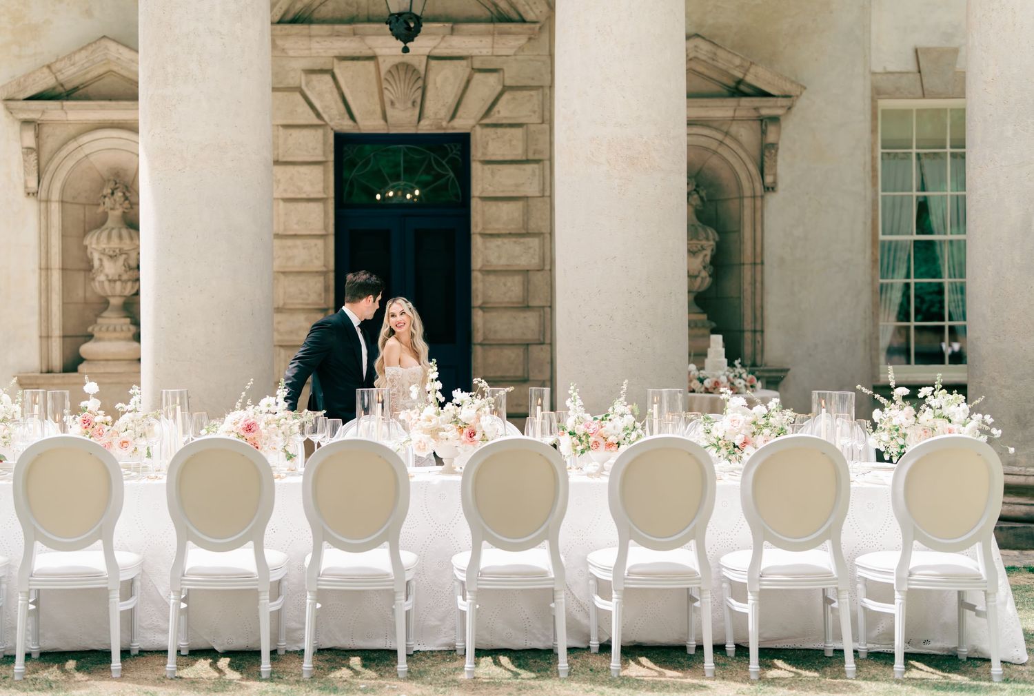 Elegant outdoor wedding reception with white Louis chairs and floral centerpieces at a grand neoclassical venue.