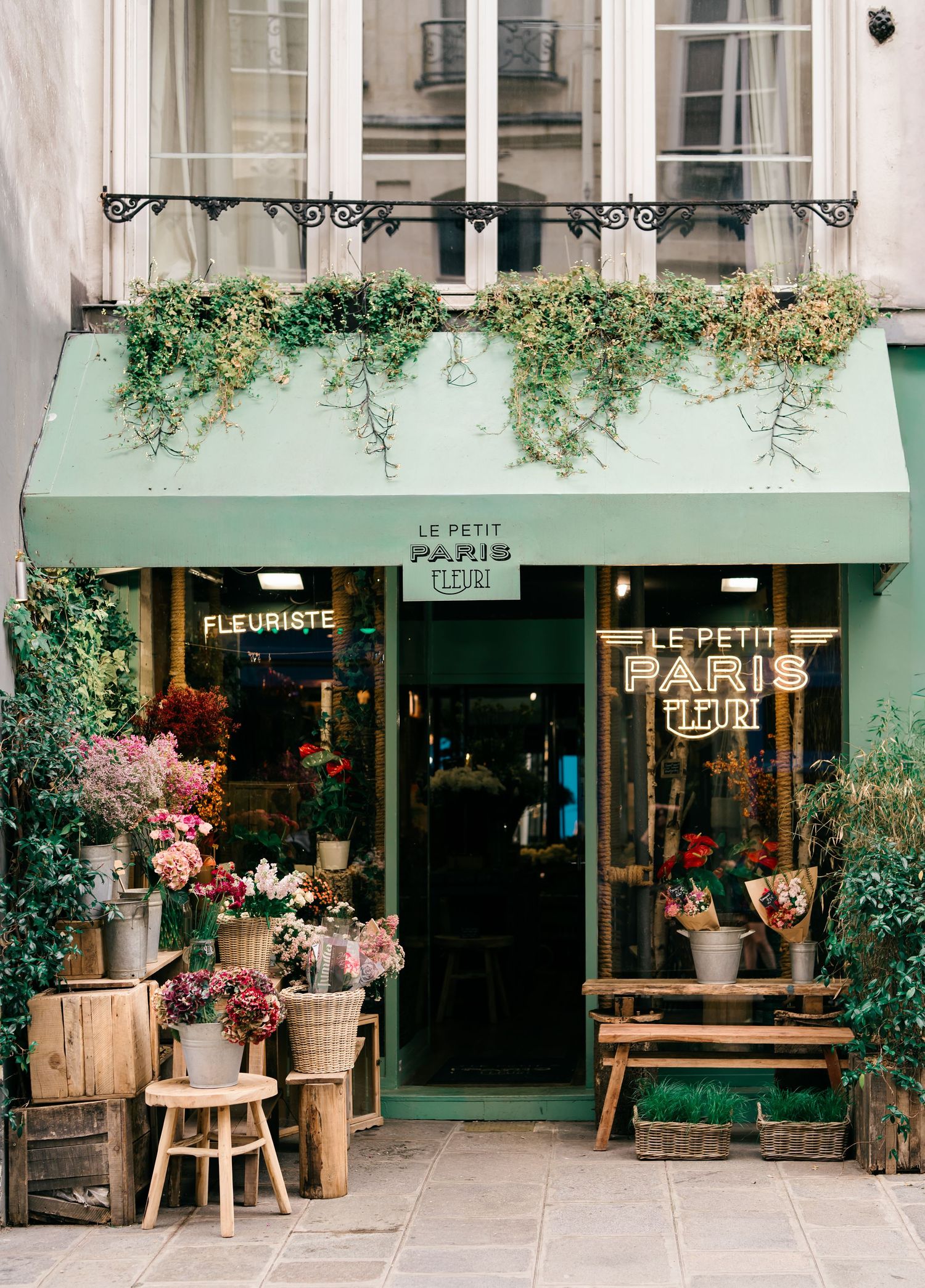 Charming Parisian flower shop Le Petit Paris with mint green facade, lush hanging plants, and colorful blooms displayed outside.