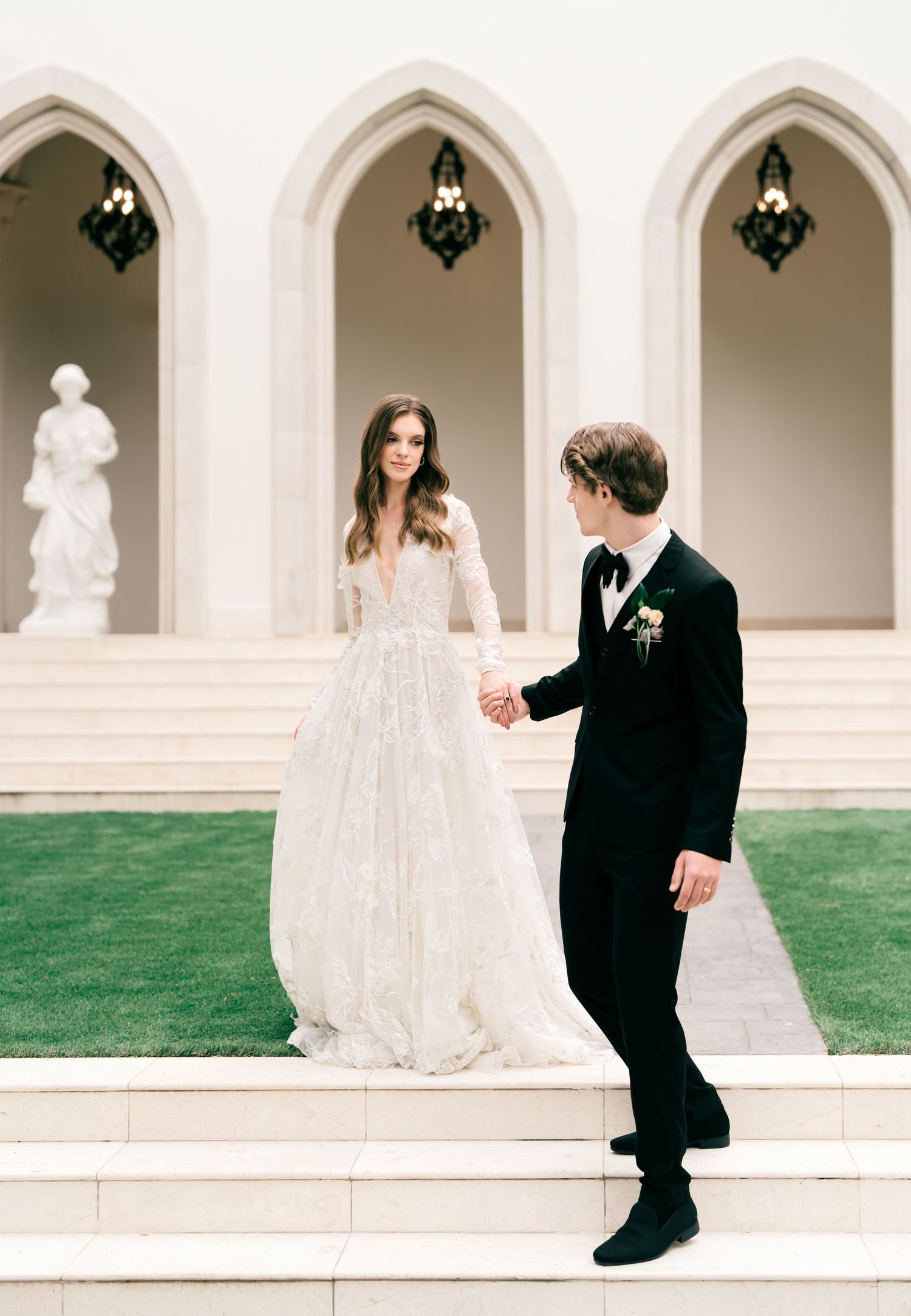 Elegant wedding couple stands on white stone steps with gothic arches and a statue in the background.