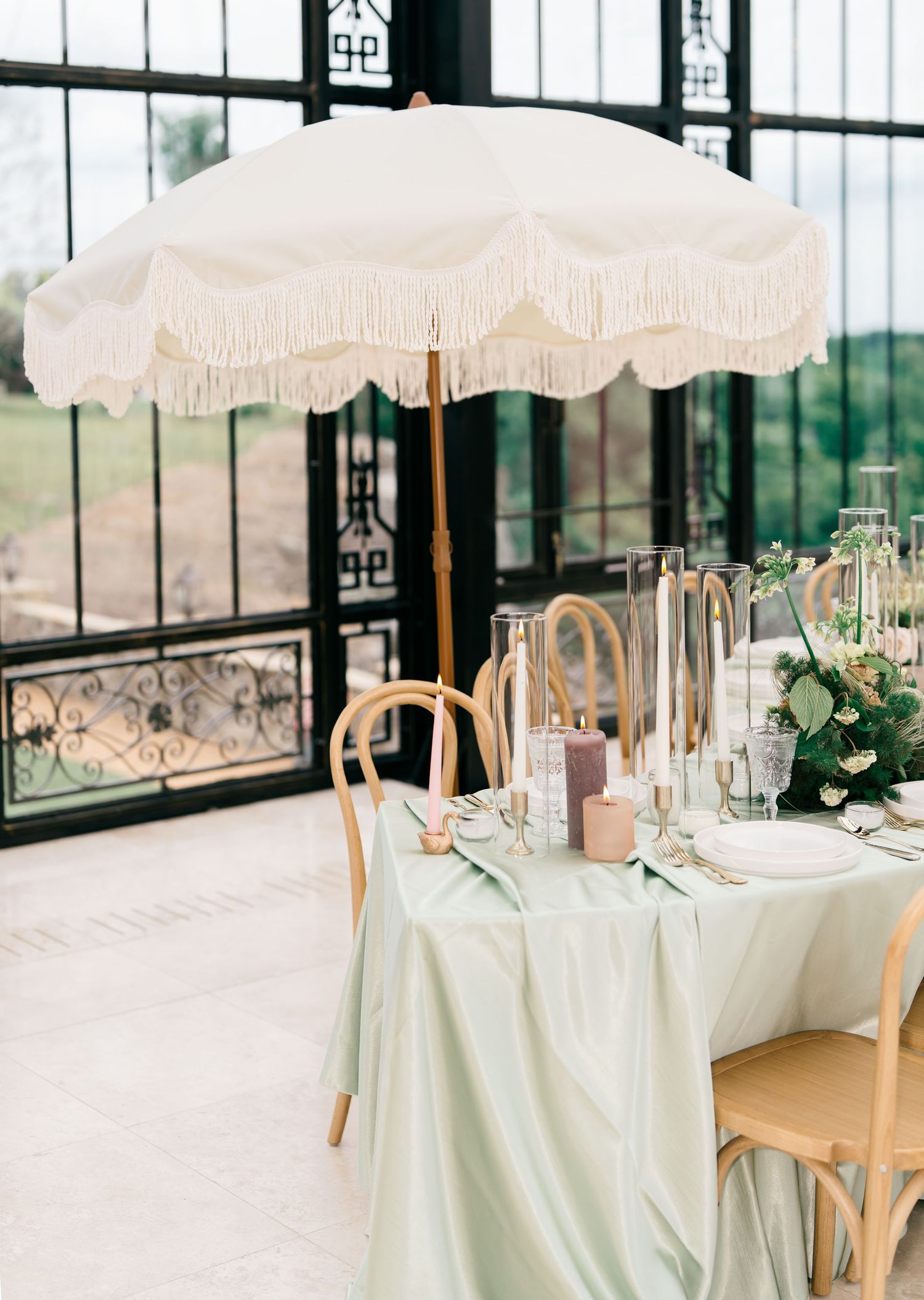 Elegant wedding reception table with mint green linen, fringed cream umbrella, candles, and floral centerpiece indoors.