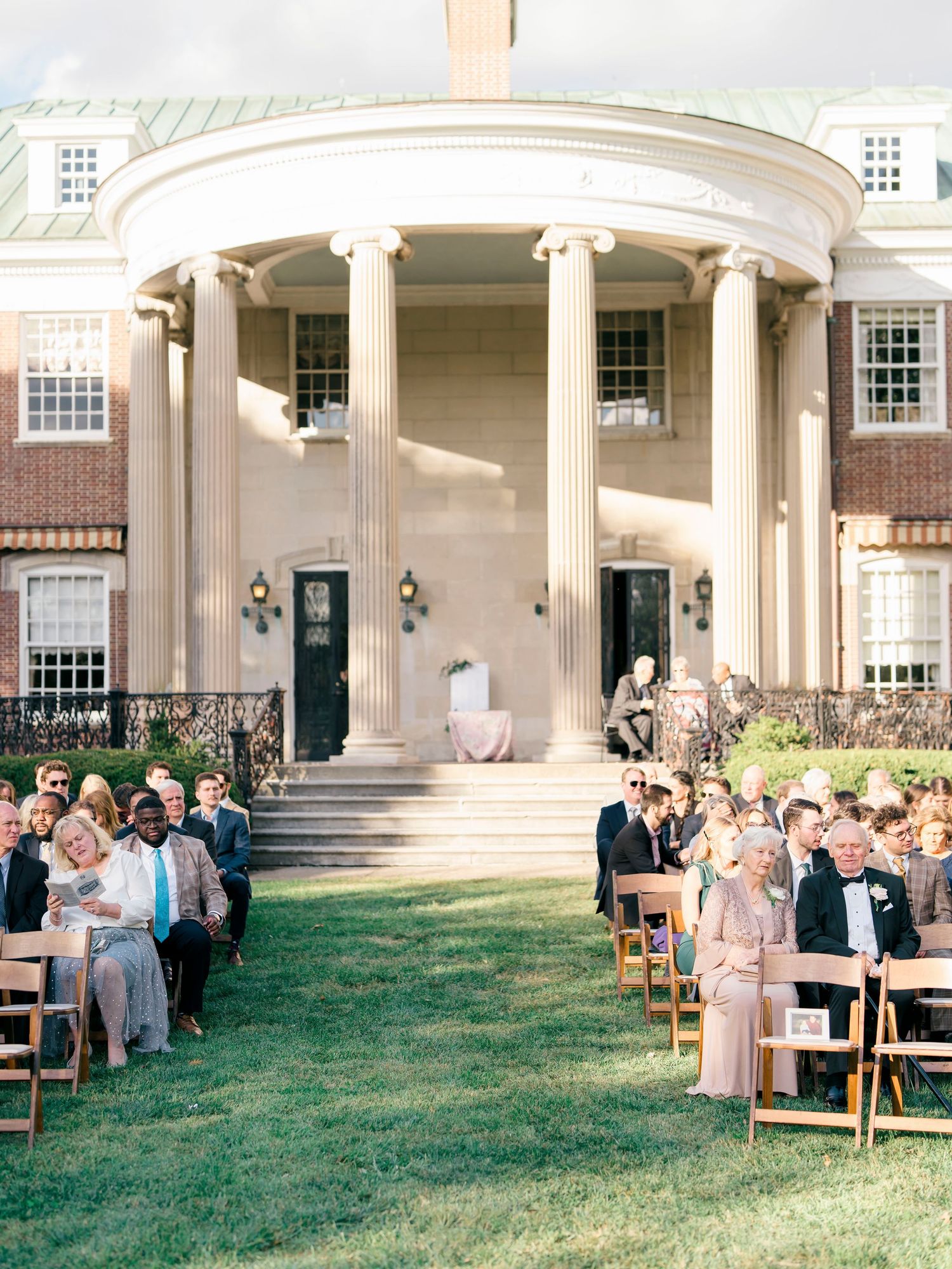 Panoramic view of an elegant outdoor wedding ceremony setup with white columns and guests seated on the lawn.