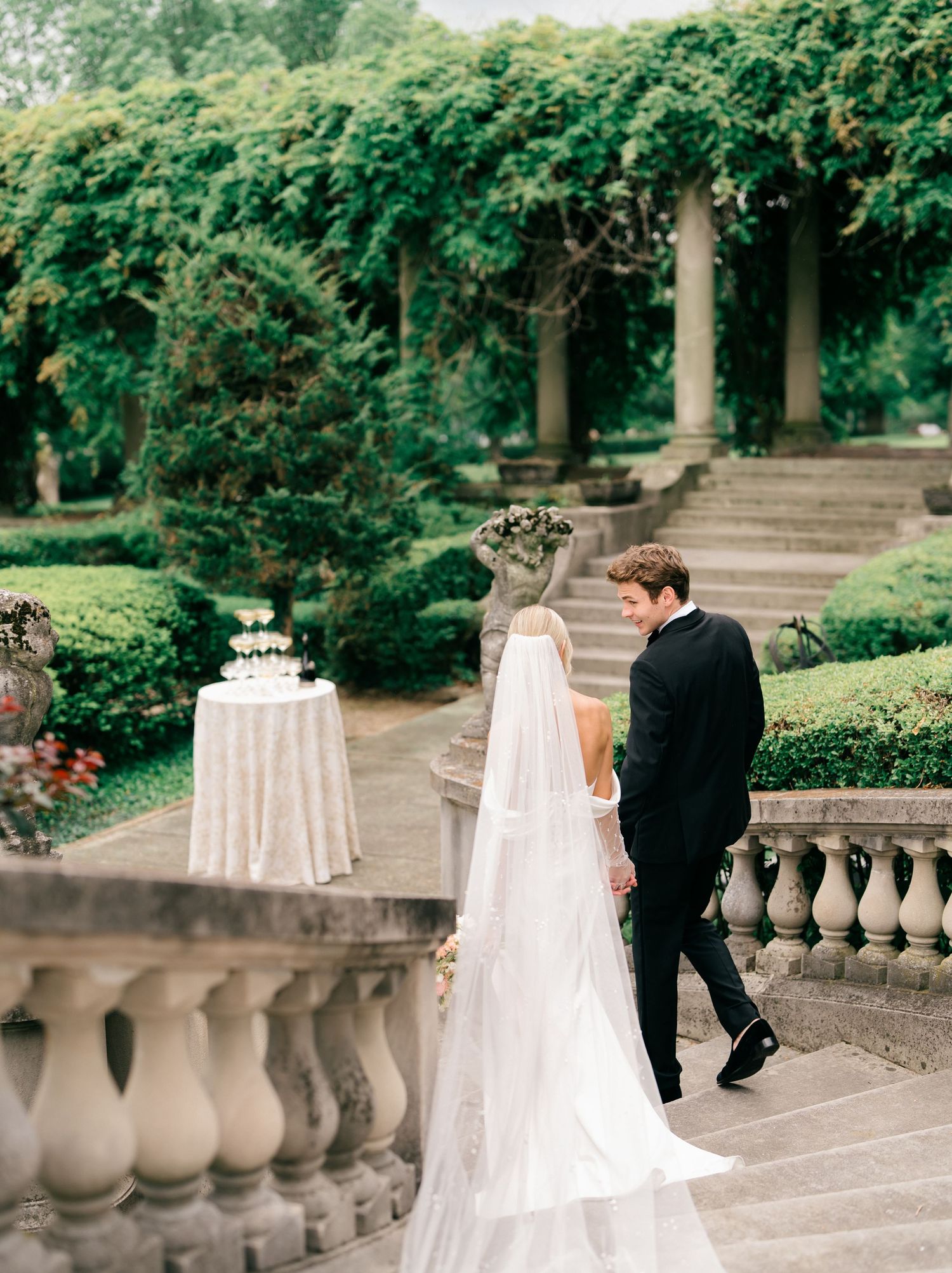 A couple walks along an elegant stone terrace surrounded by lush greenery in a romantic garden setting.