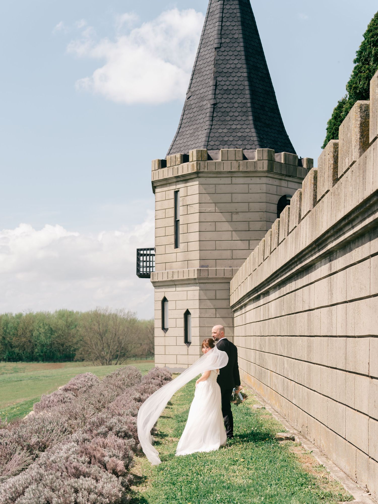 At The Kentucky Castle. Bride and groom kissing beside a stone castle tower with a black spire, veil blowing in the wind on a sunny day.