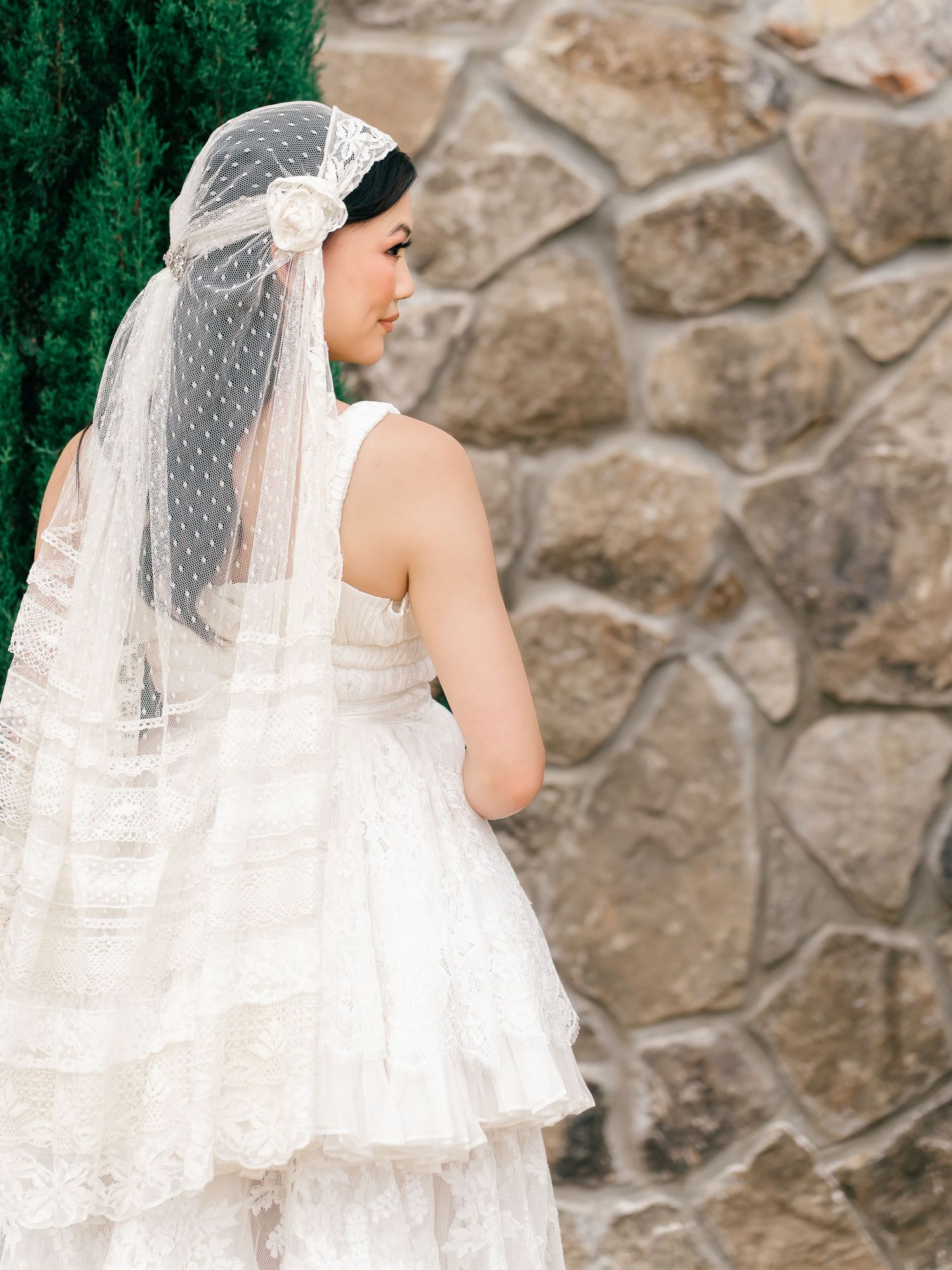 Bride in layered lace wedding dress and dotted birdcage veil with floral accent, posed against rustic stone wall.