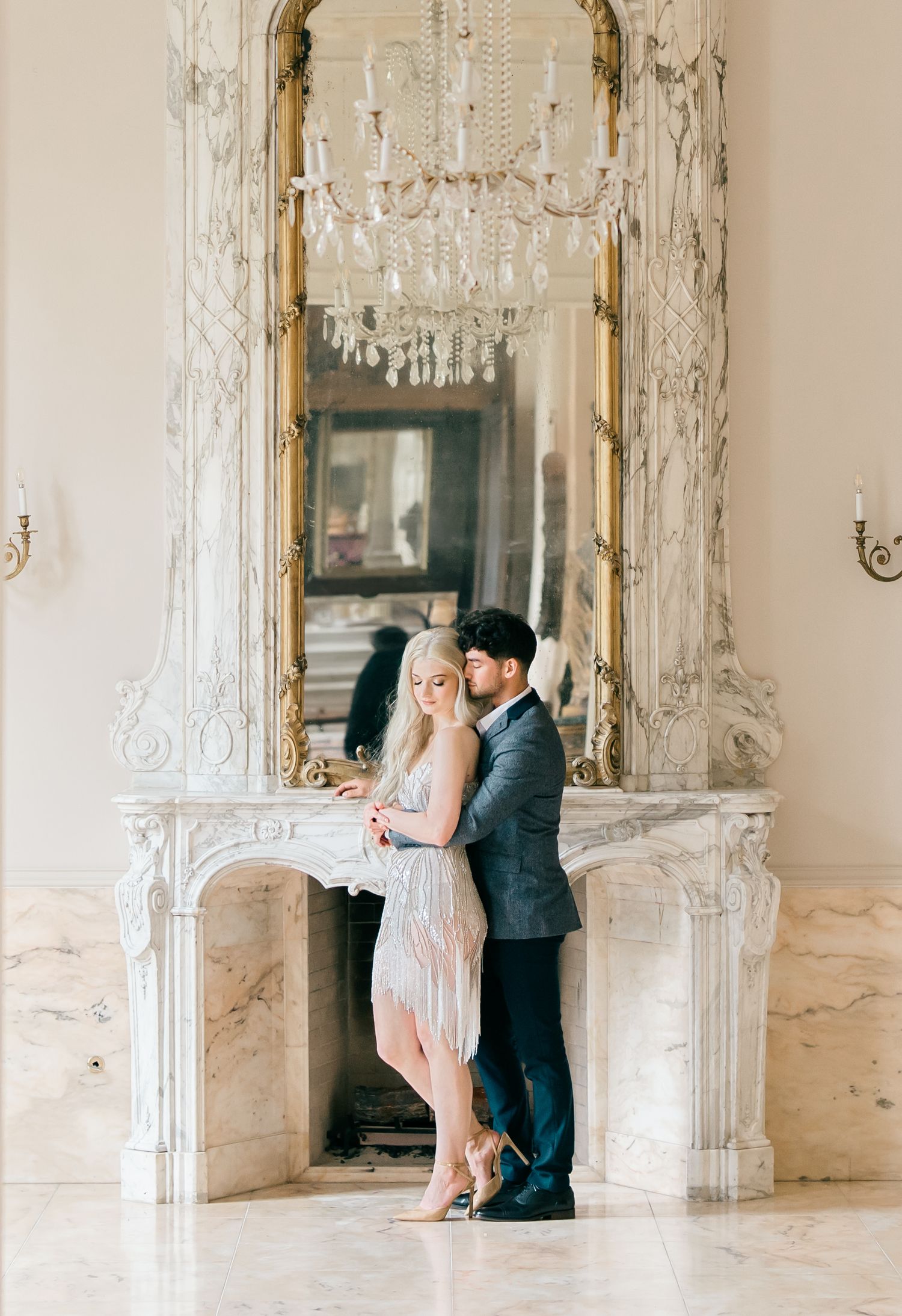 A romantic couple poses together in an elegant white room with ornate mirrors and crystal chandeliers.