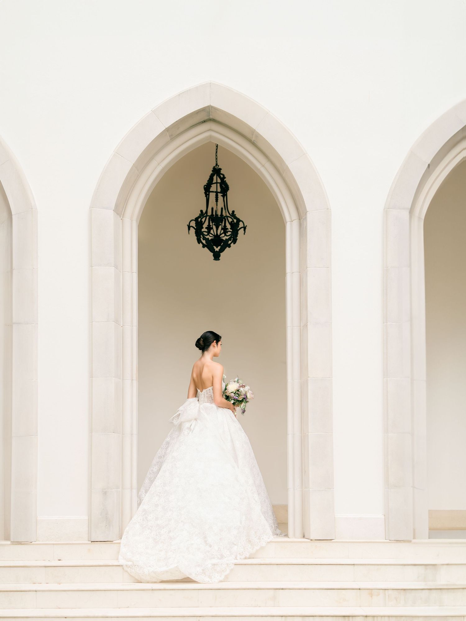 Elegant bride in flowing white gown poses in a series of arched white hallways with hanging chandeliers.