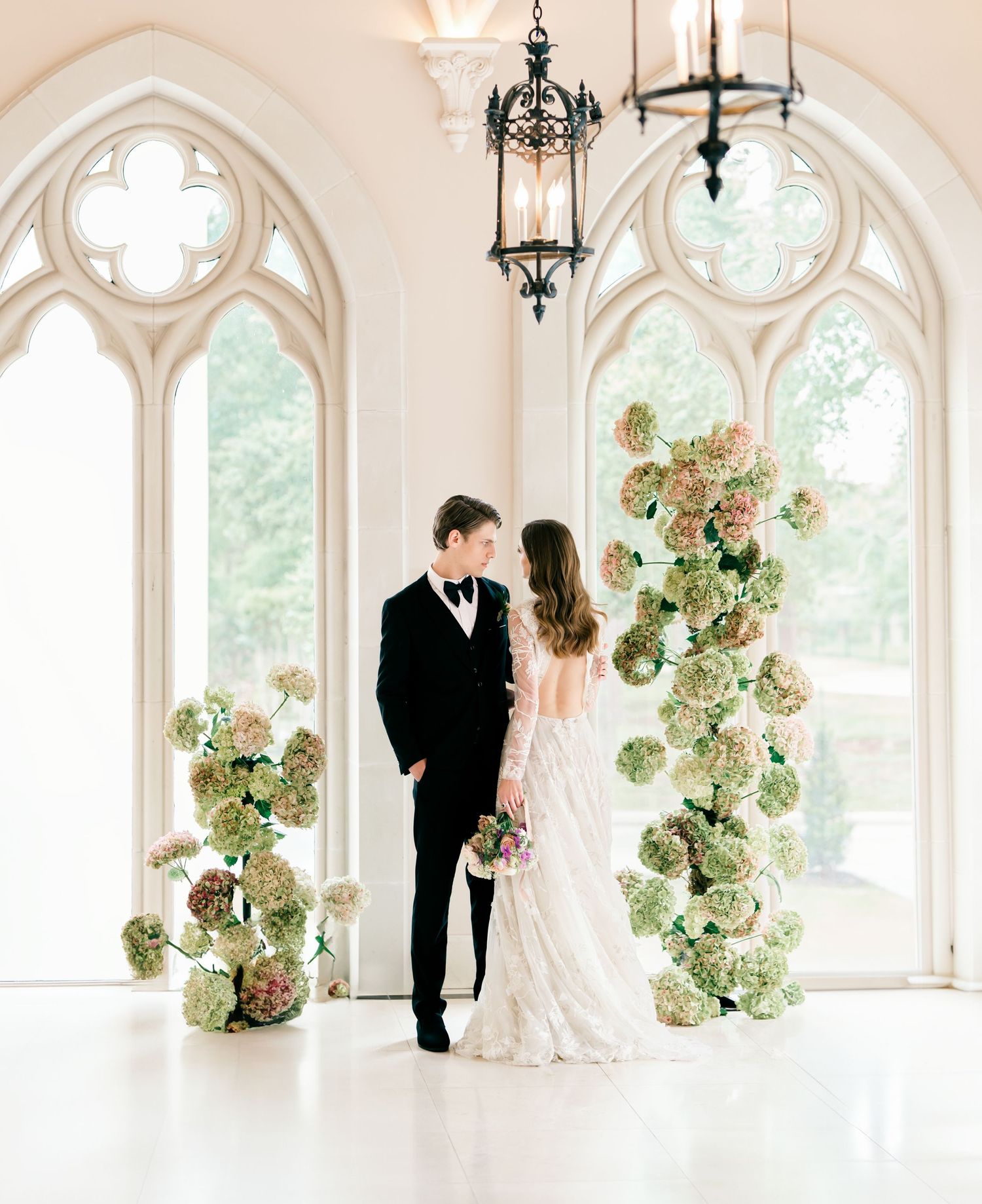 Elegant wedding scene in a bright chapel with Gothic windows and cascading green hydrangea floral arrangements.