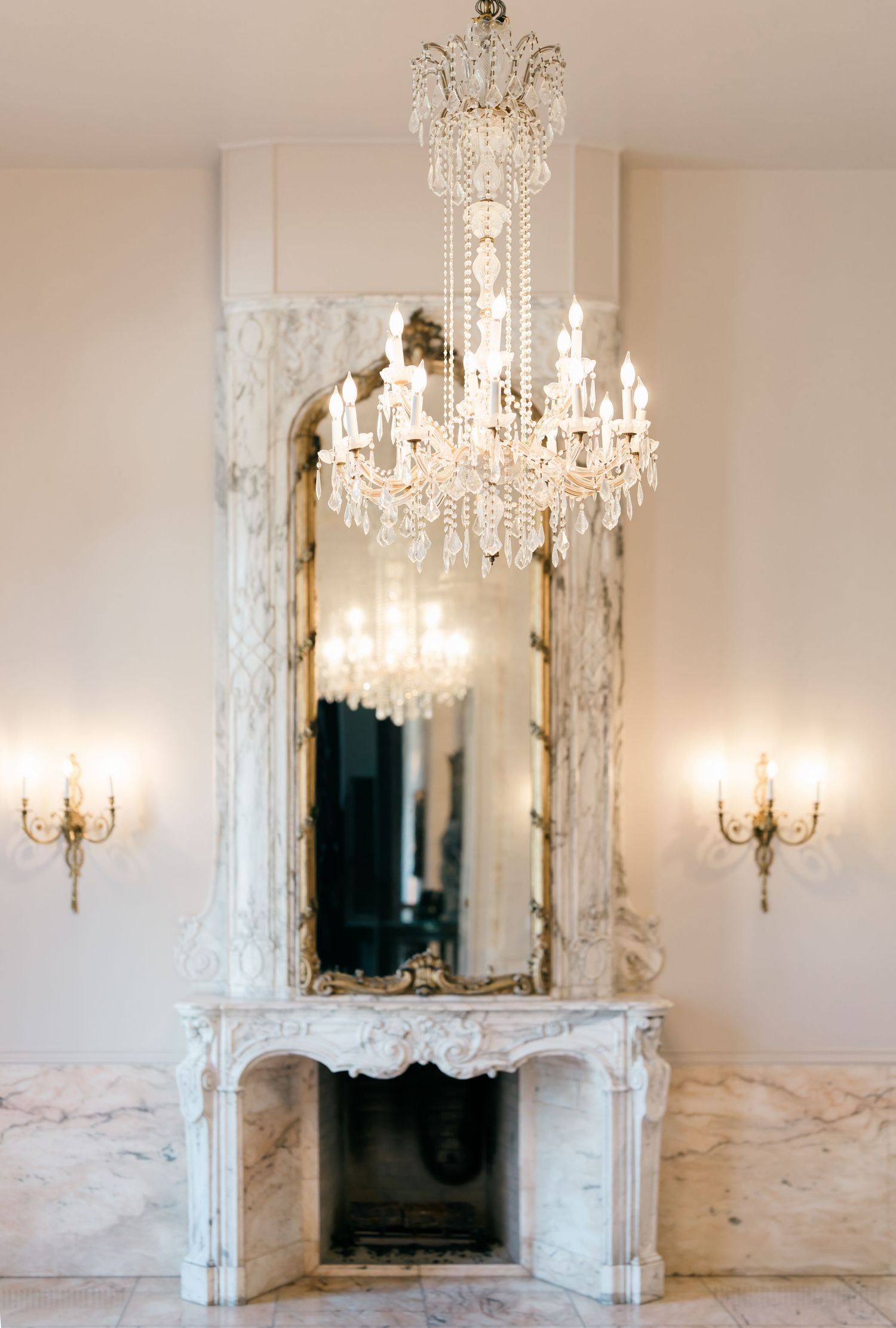 Elegant white marble fireplace with ornate crystal chandelier in a luxurious room setting.