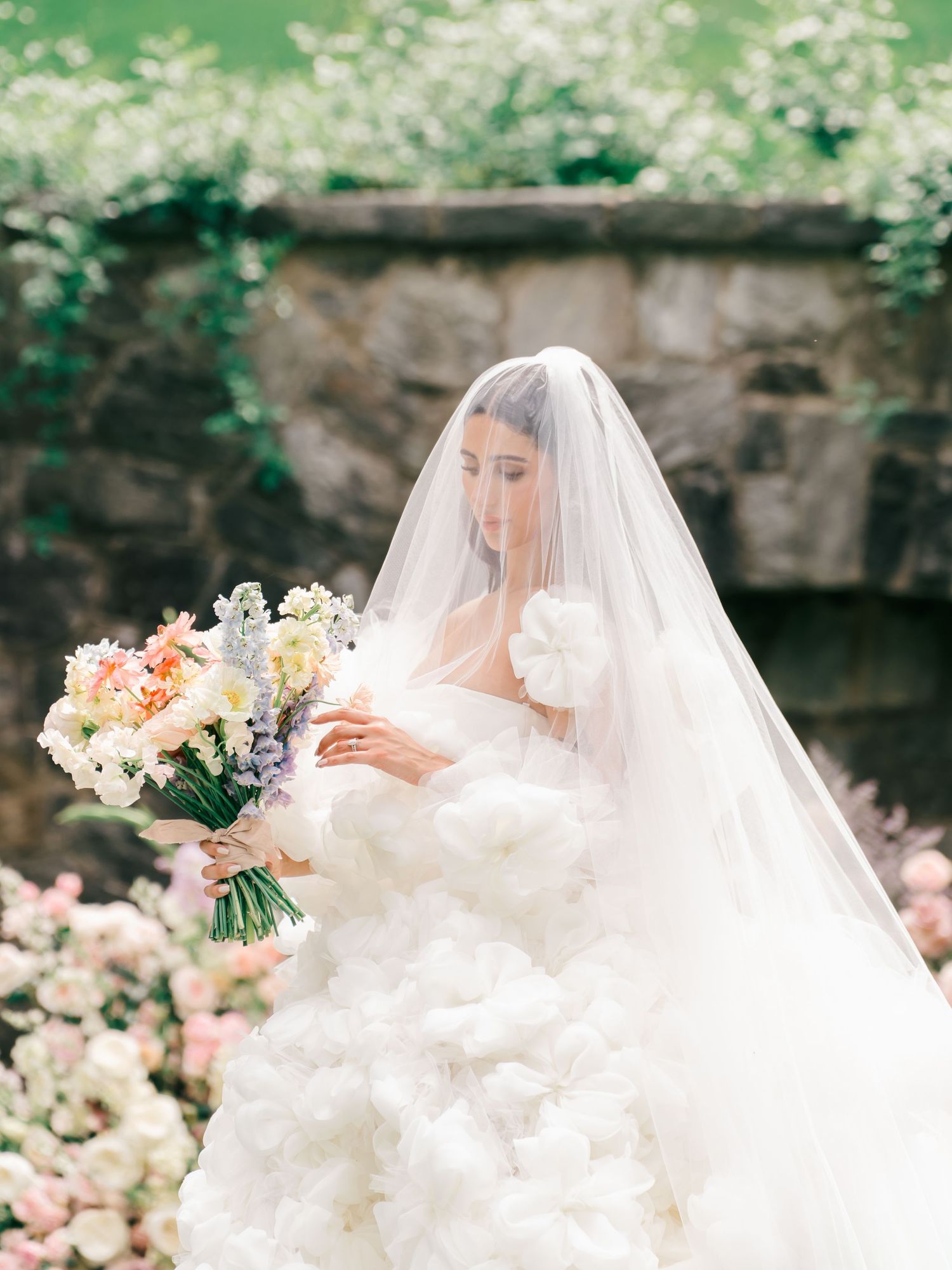 Bride in a voluminous white ruffled gown and veil holding a pastel bouquet in a lush garden setting.