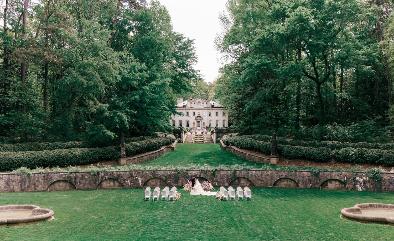 Elegant estate wedding ceremony on lush green lawn with bridal party, surrounded by manicured hedges and towering trees.