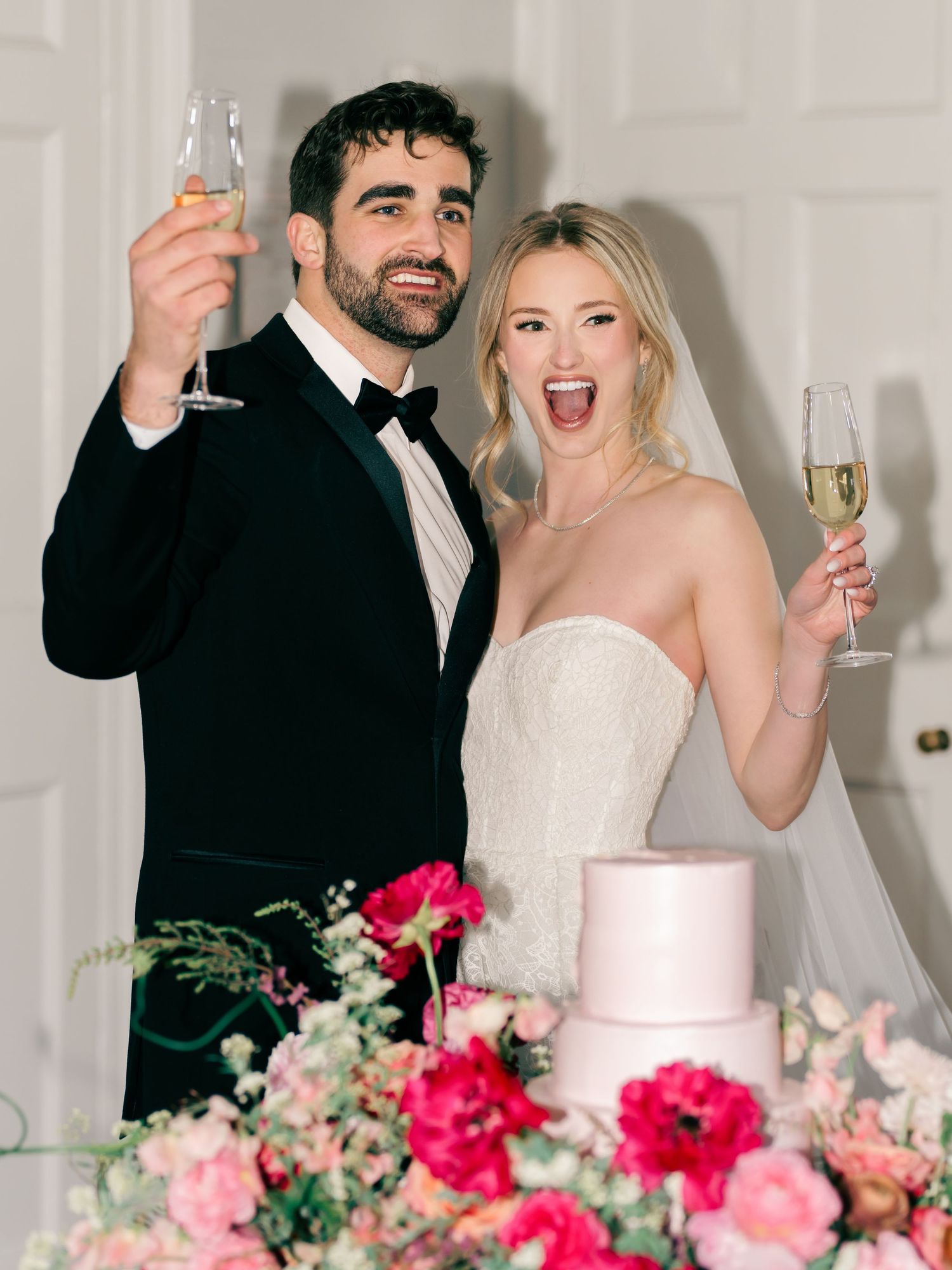 Newlyweds toast with champagne glasses beside an elegant pink wedding cake adorned with vibrant pink floral arrangements.