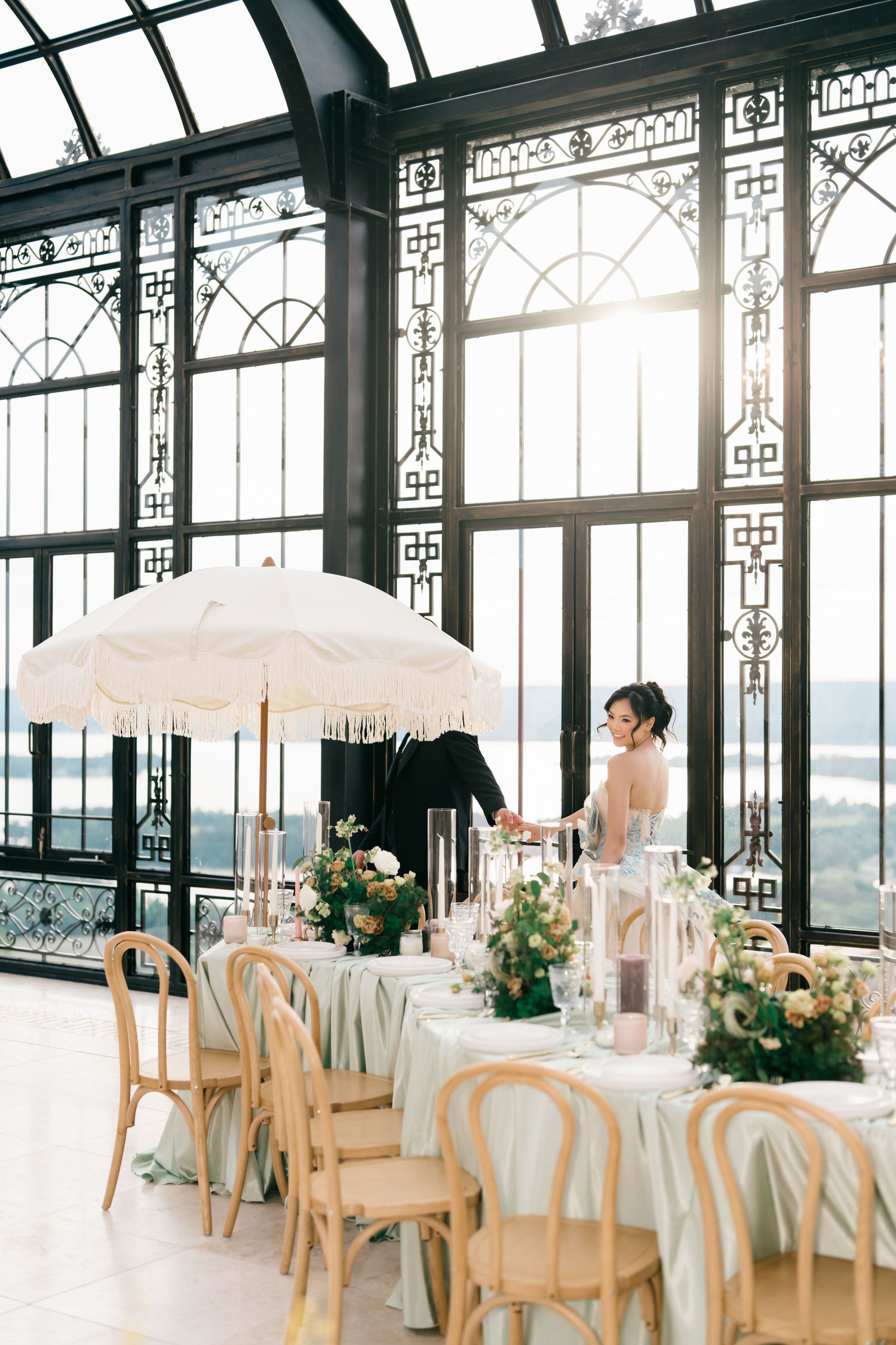 Elegant wedding reception in a sunlit conservatory with floral centerpieces, candles, a fringed umbrella, and wooden chairs.