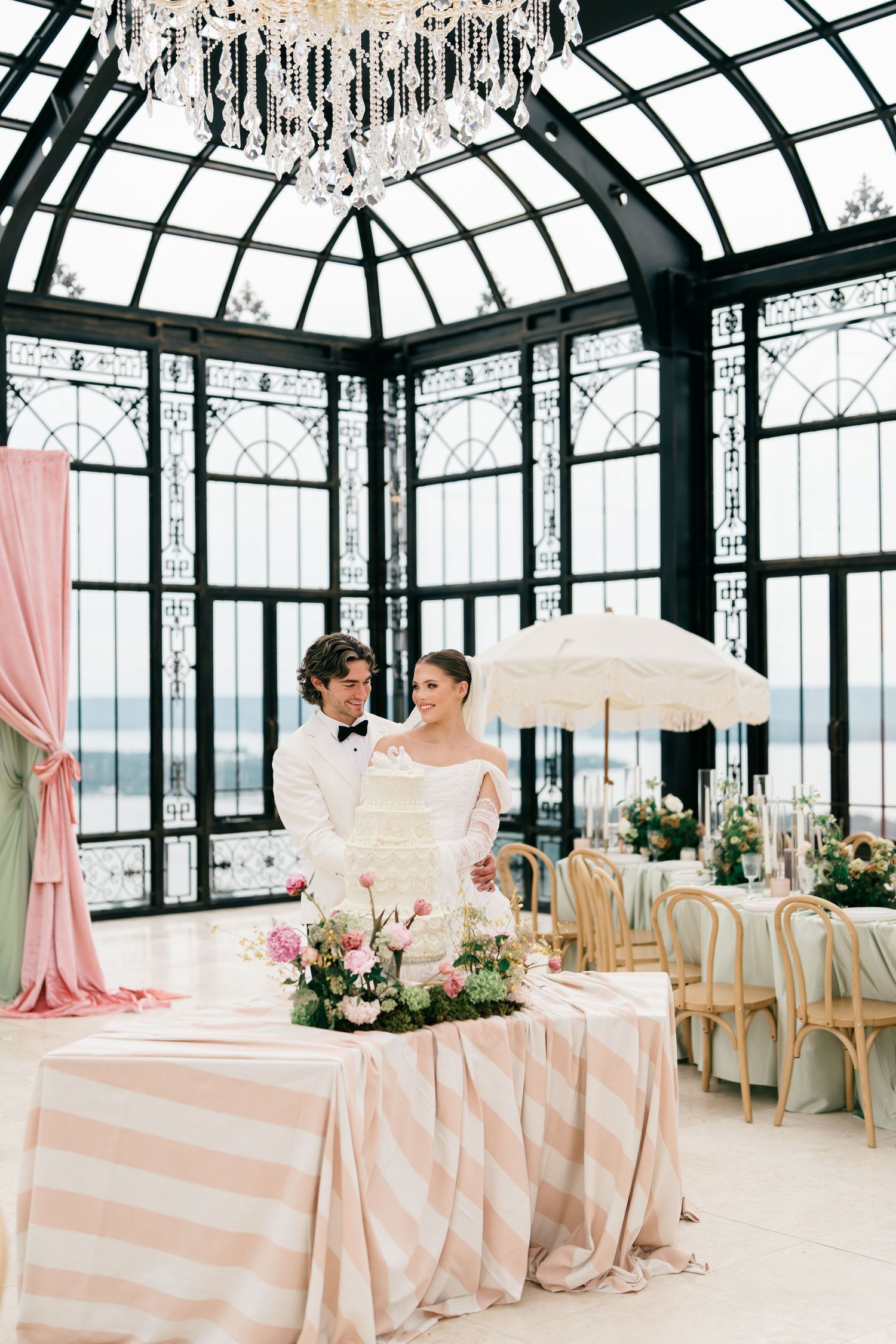 Bride and groom stand together in an elegant glass conservatory wedding venue with pink striped tablecloths and crystal chandelier.