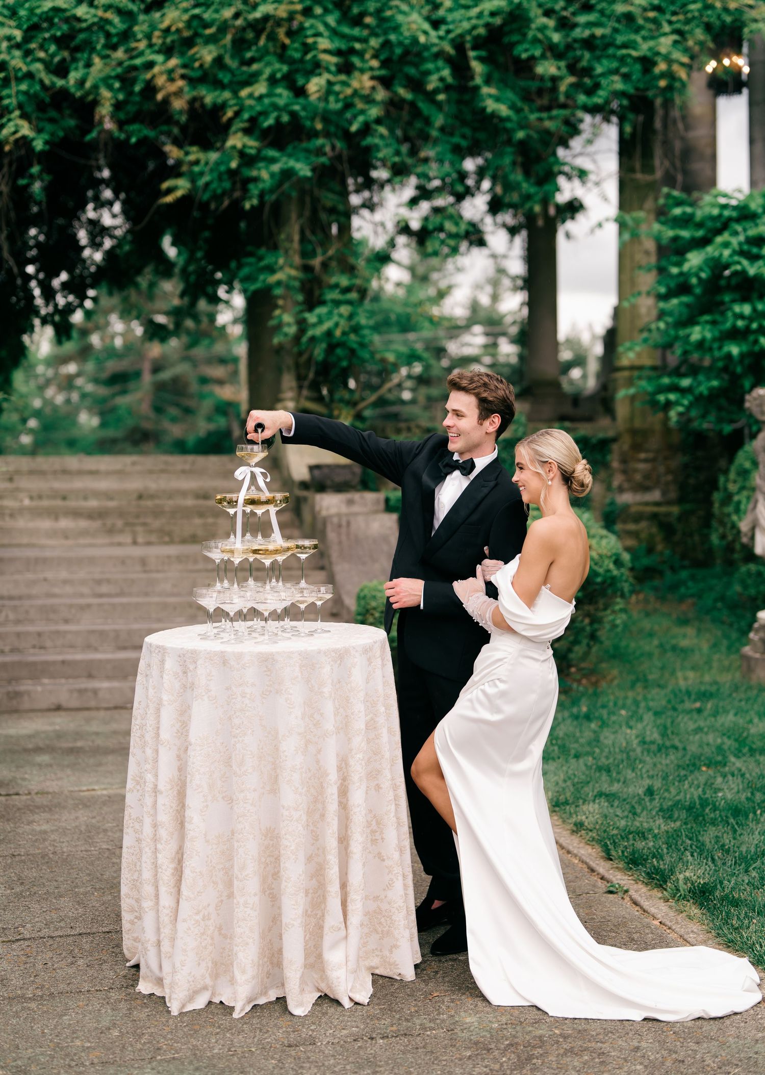 A wedding couple pours champagne into a tower of elegant crystal glasses on a lace-covered table in an outdoor garden setting.