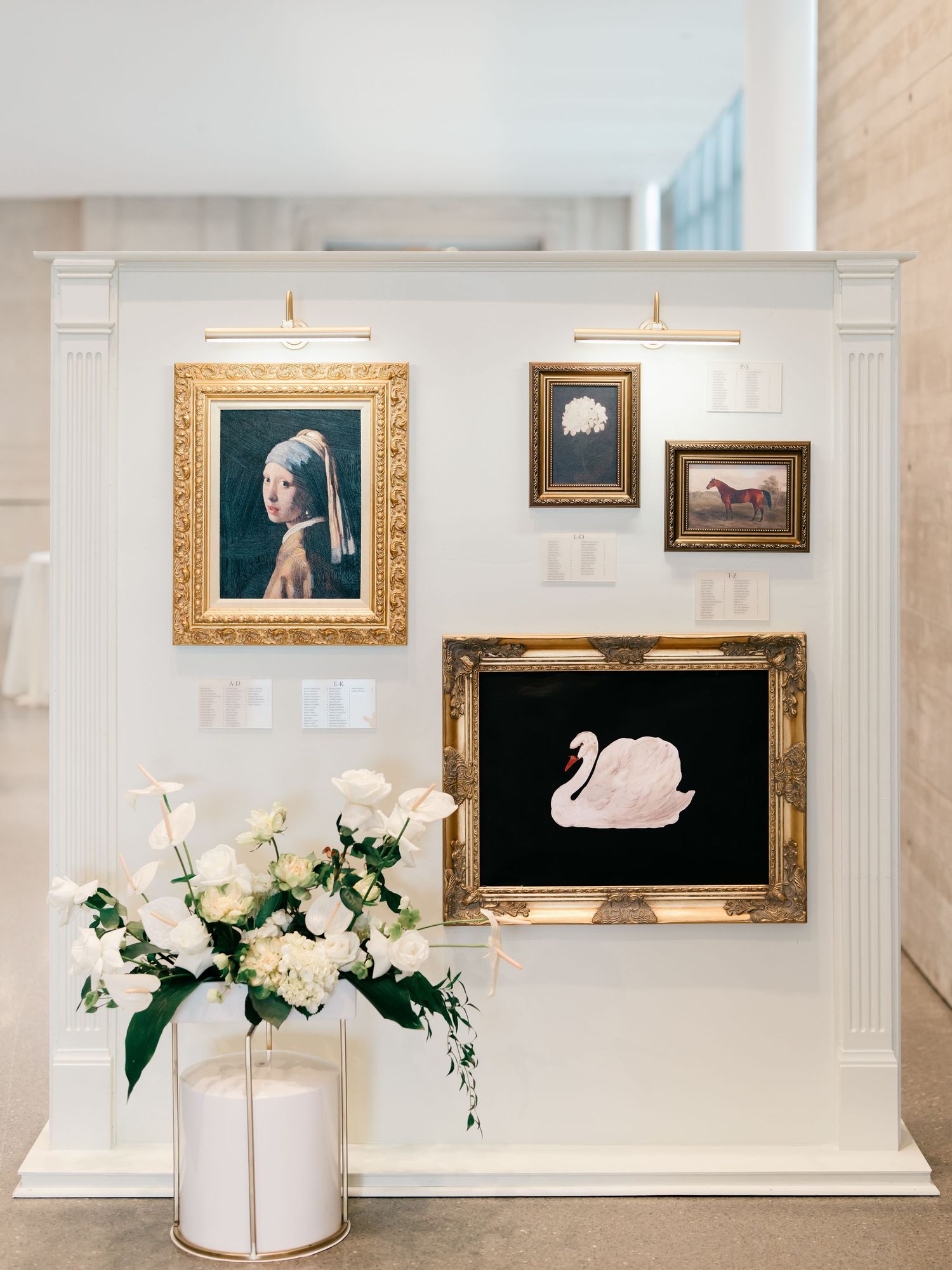 Elegant gallery wall display with ornate gold frames featuring classic artworks and a white floral arrangement in a modern space.