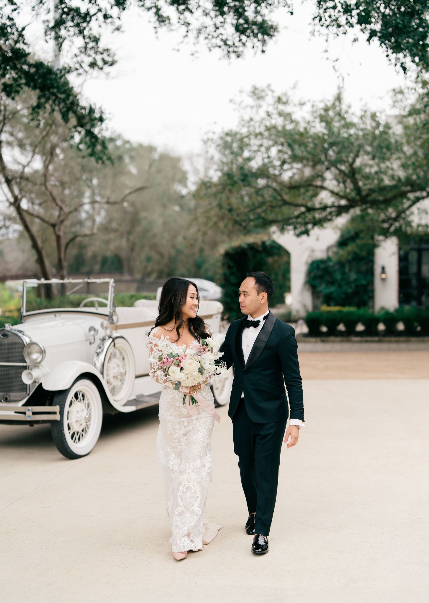 Elegant couple dancing and twirling outdoors in formal attire next to vintage white car in garden setting.