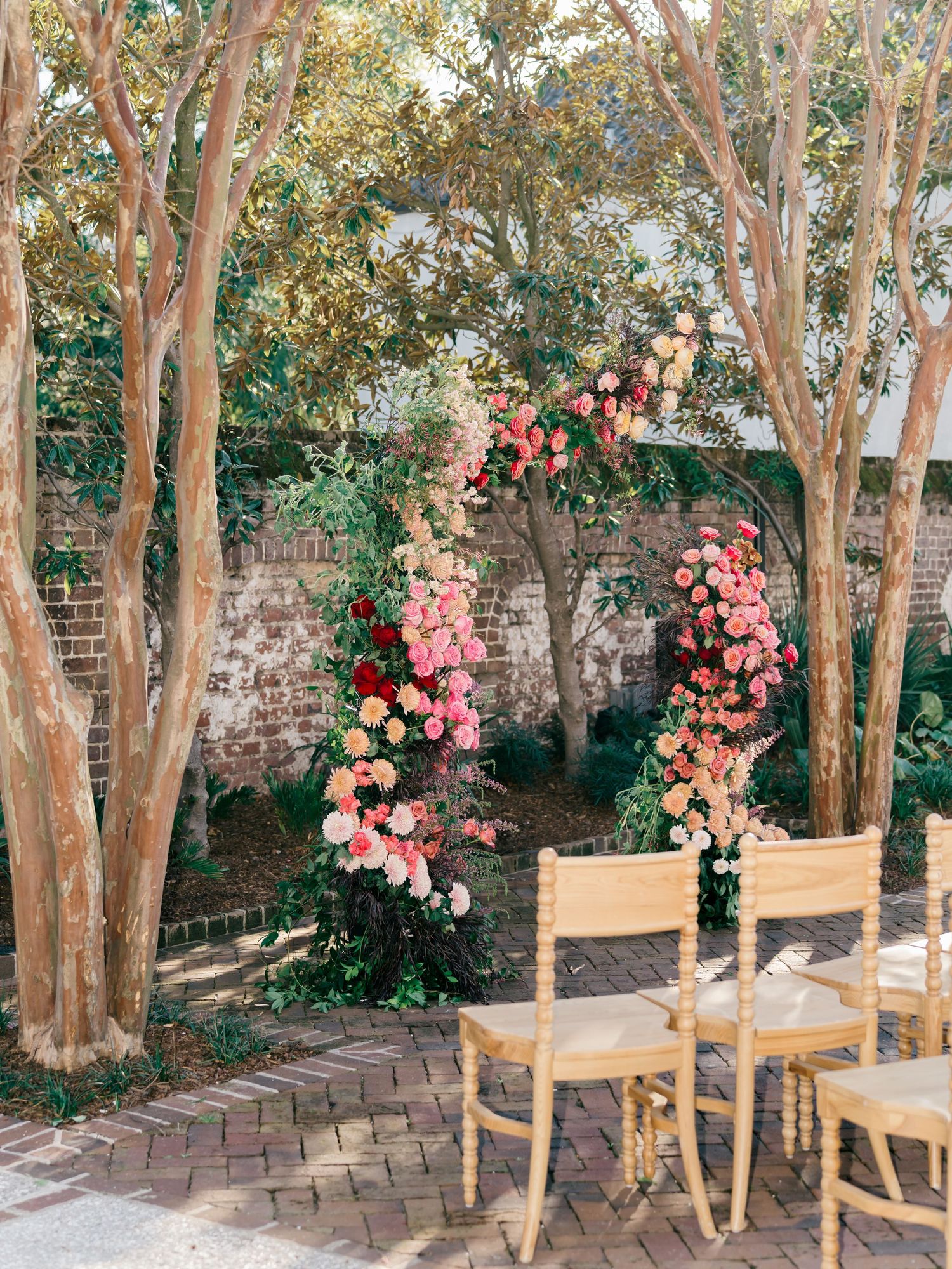 Wooden chairs arranged in a garden setting with pink floral arrangements and crepe myrtle trees creating a romantic atmosphere.