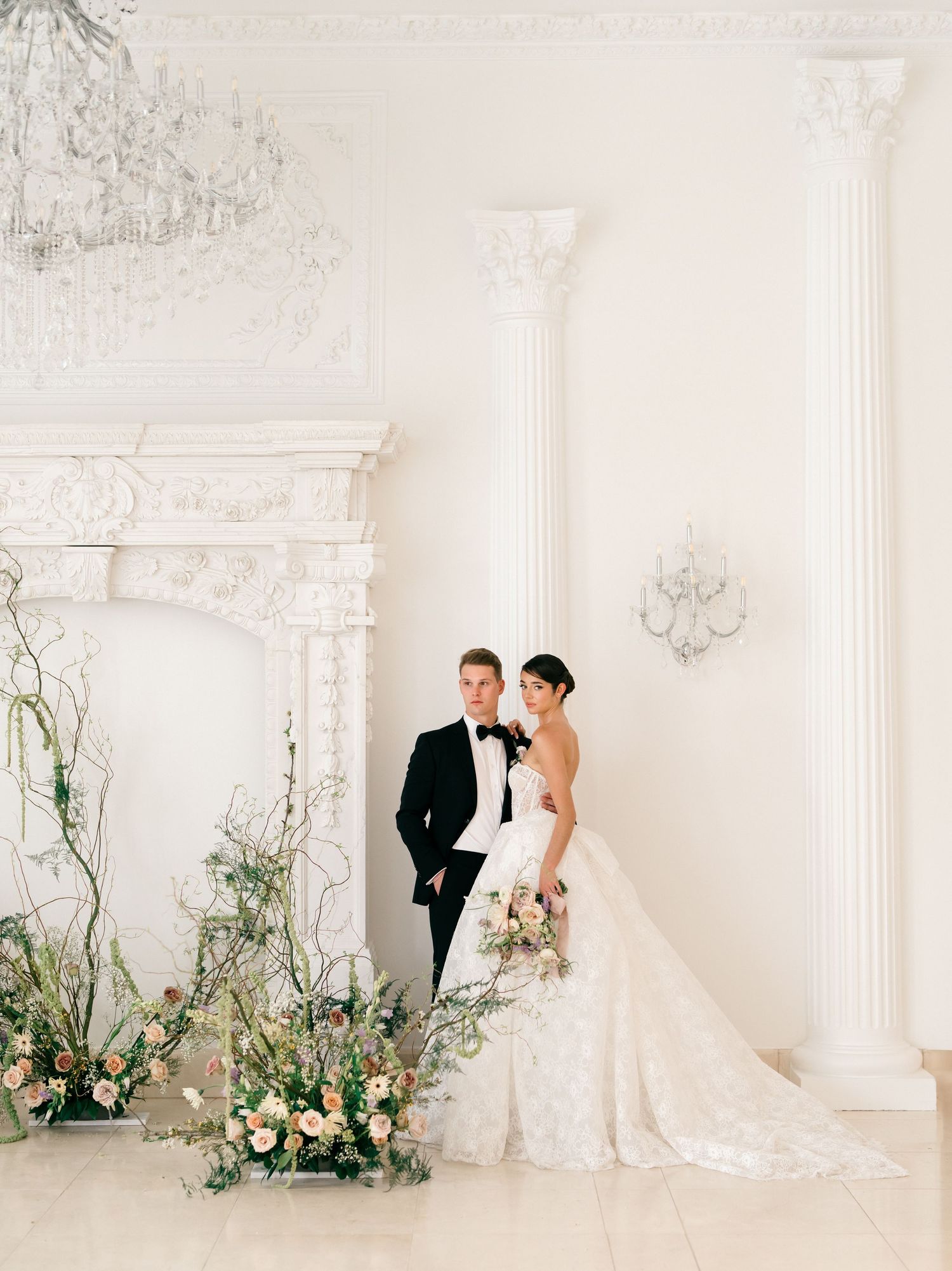 Elegant wedding couple poses in a bright white room with ornate architectural details and floral arrangements.