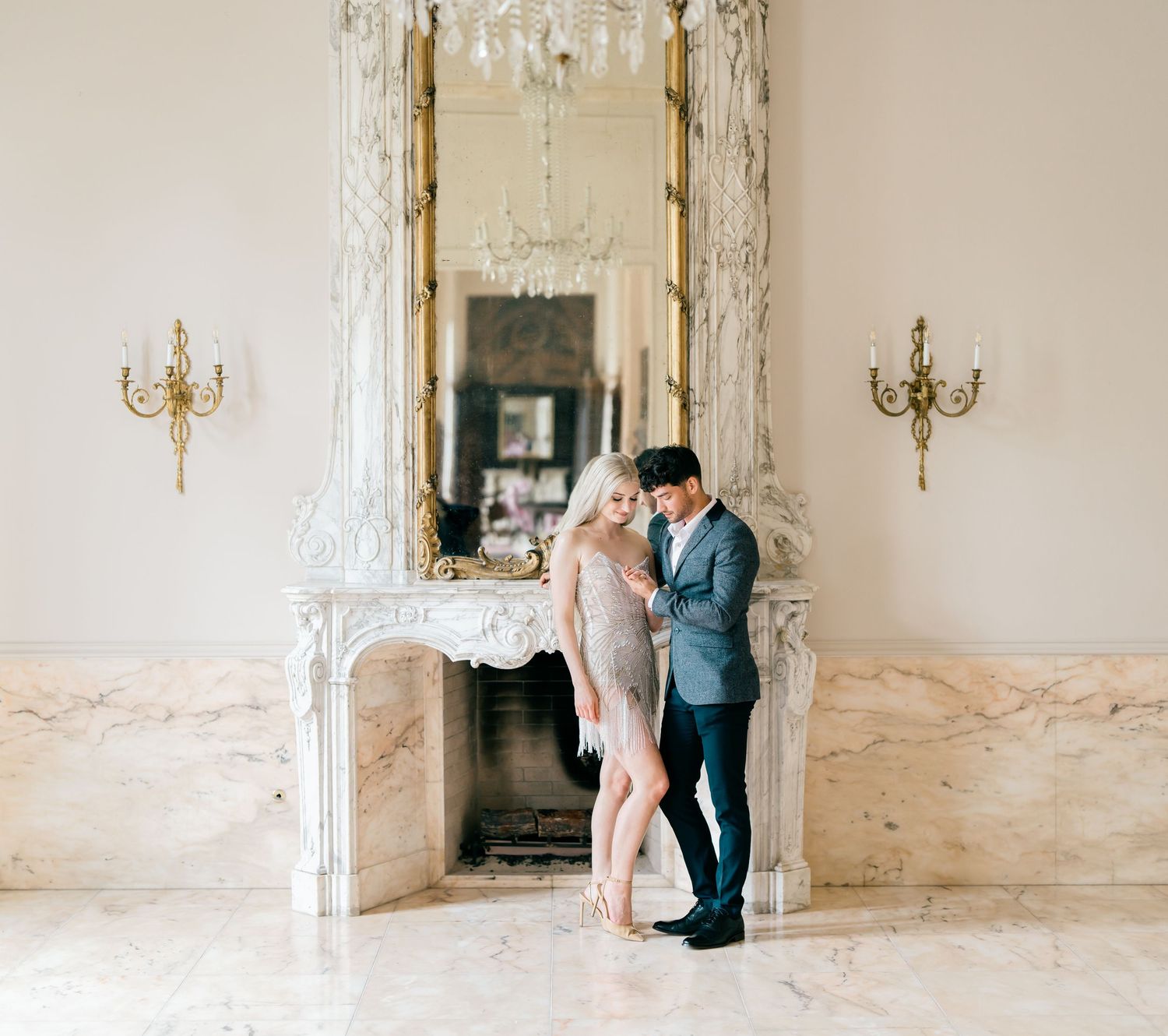 A couple embraces in a luxurious white marble ballroom with crystal chandeliers and gold wall sconces.