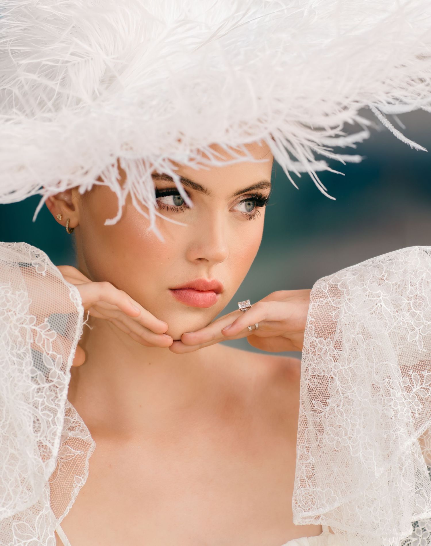 Elegant bride wearing a wide-brimmed white feathered hat and lace veil, posing gracefully with hands under chin.