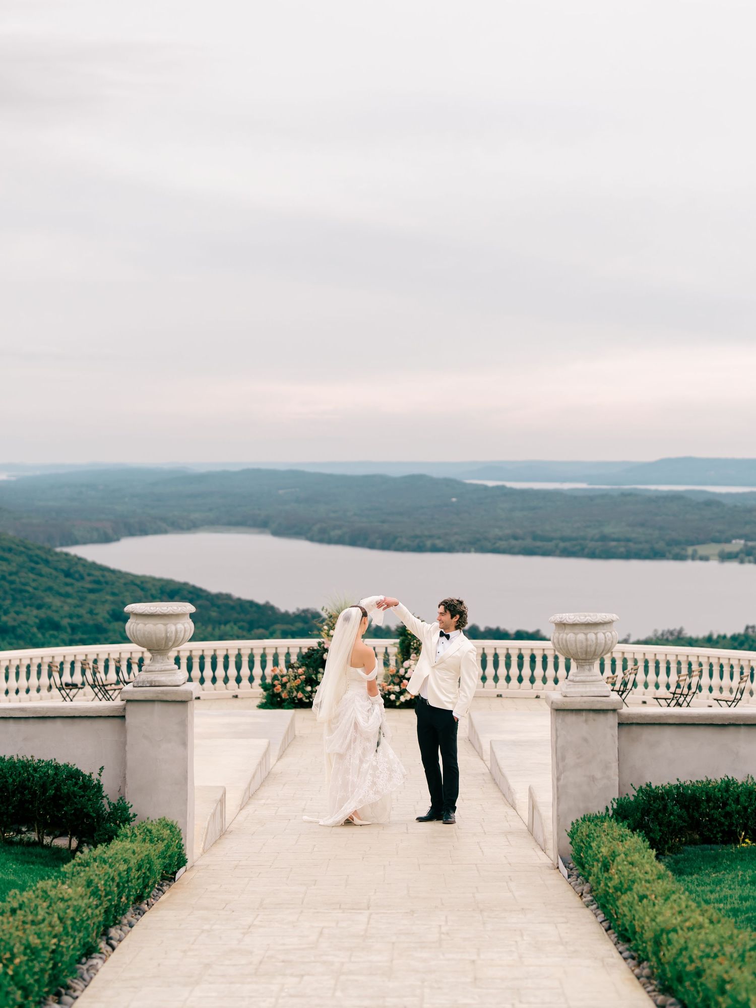 Bride and groom share a romantic moment on an elegant terrace overlooking a scenic lake and rolling hills.