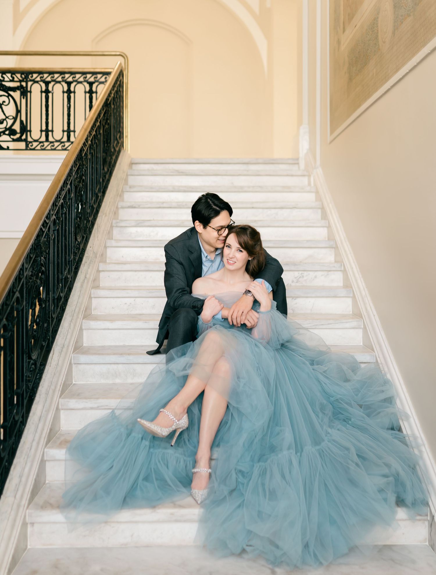 A romantic couple in formal attire poses on elegant white stairs, with the bride wearing a flowing dusty blue tulle gown.