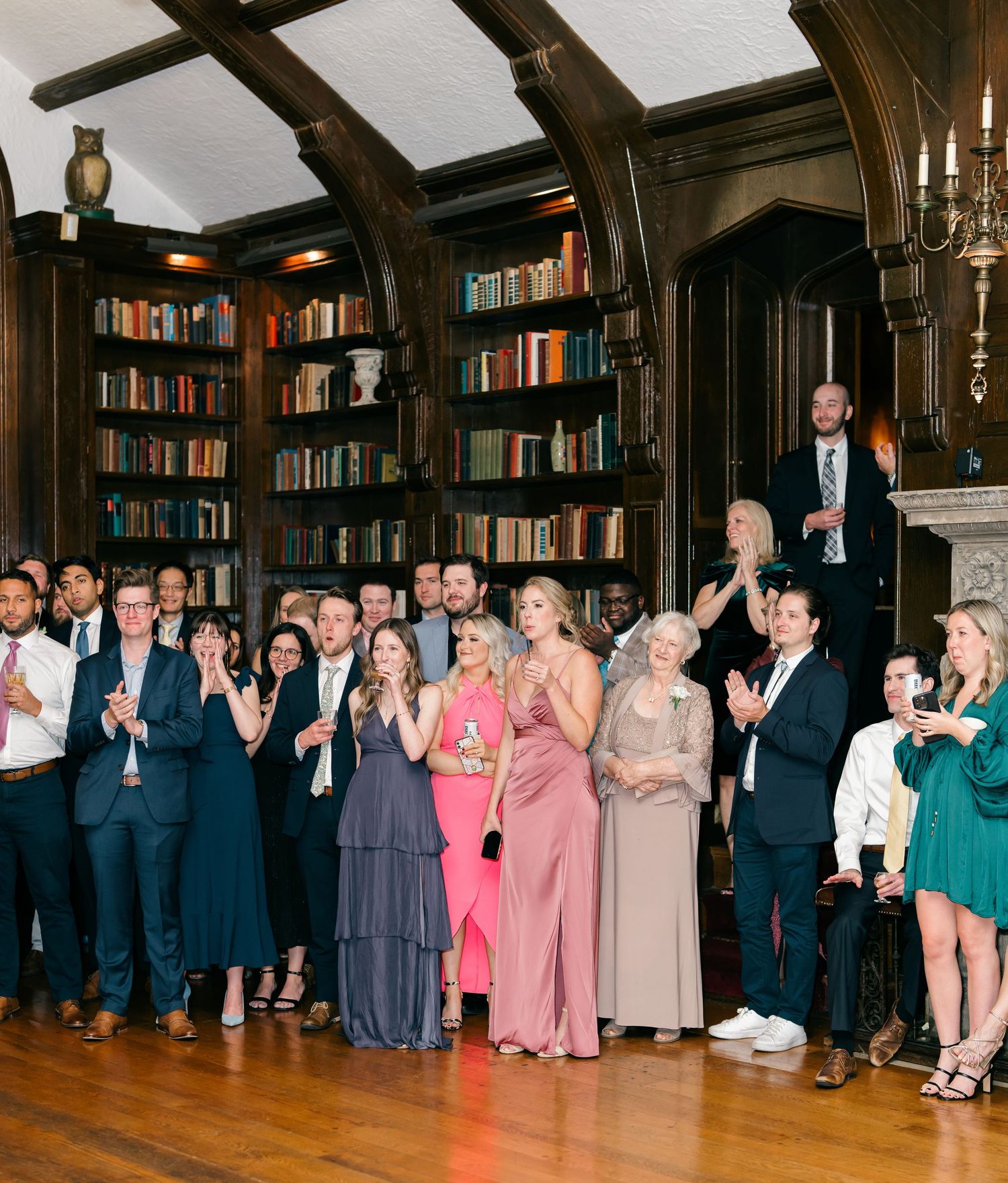 Wedding guests in formal attire dance and celebrate together in an elegant wood-paneled library room with bookshelves.