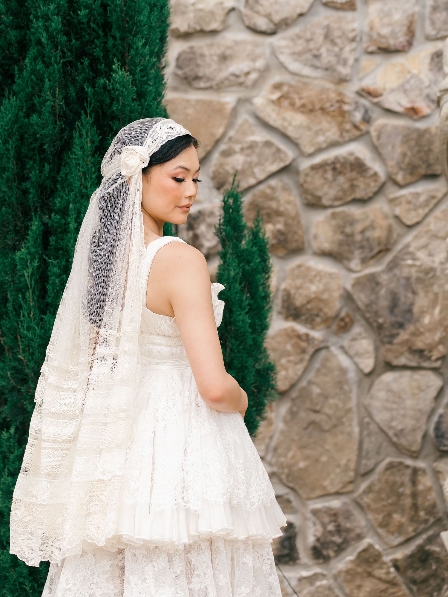 Bride in layered lace wedding dress and dotted chapel veil with floral headpiece stands by stone wall.