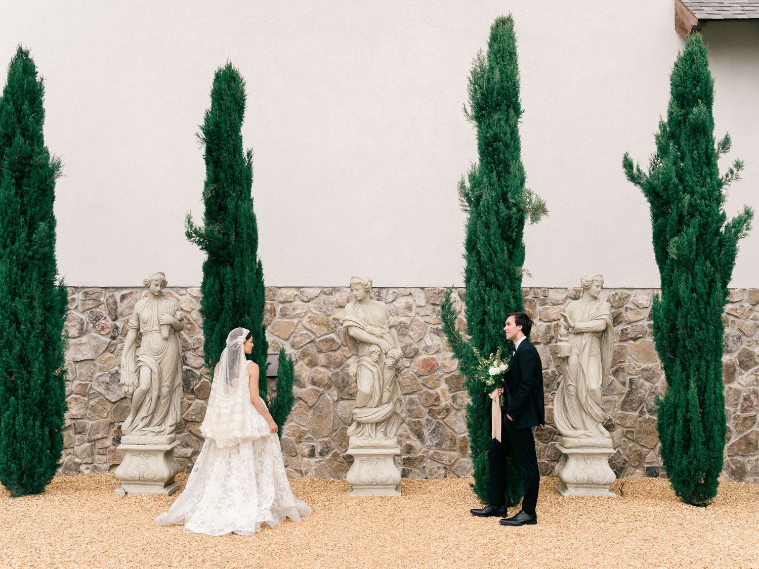 Bride and groom stand among stone statues and tall cypress trees at an elegant outdoor wedding venue.