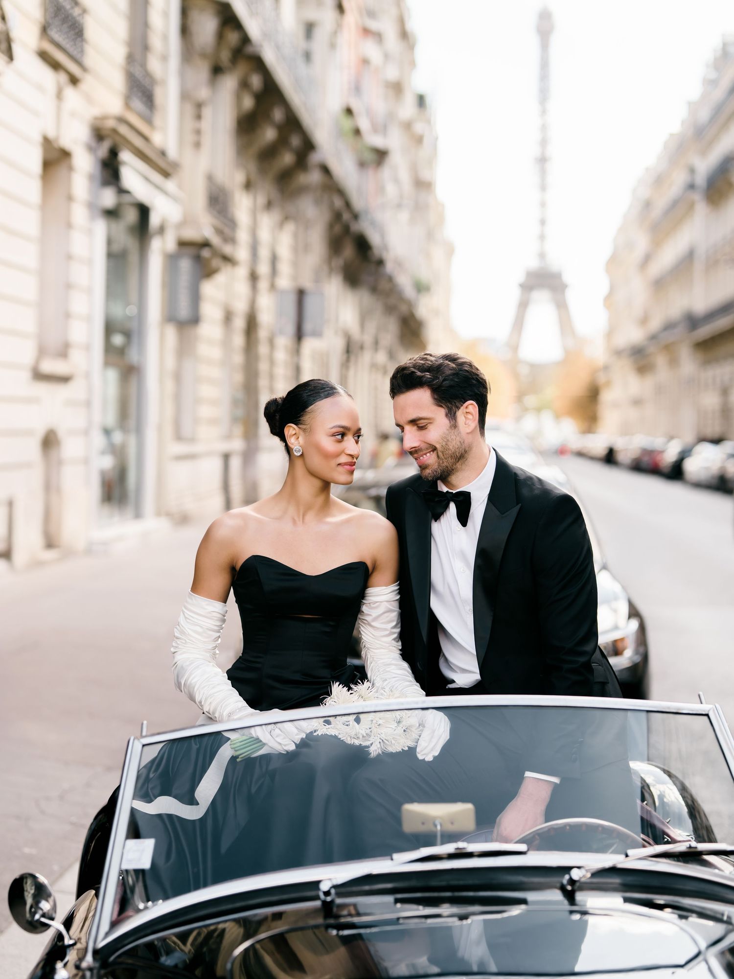 Romantic couple in formal attire sharing intimate moments in a vintage convertible car with the Eiffel Tower visible.