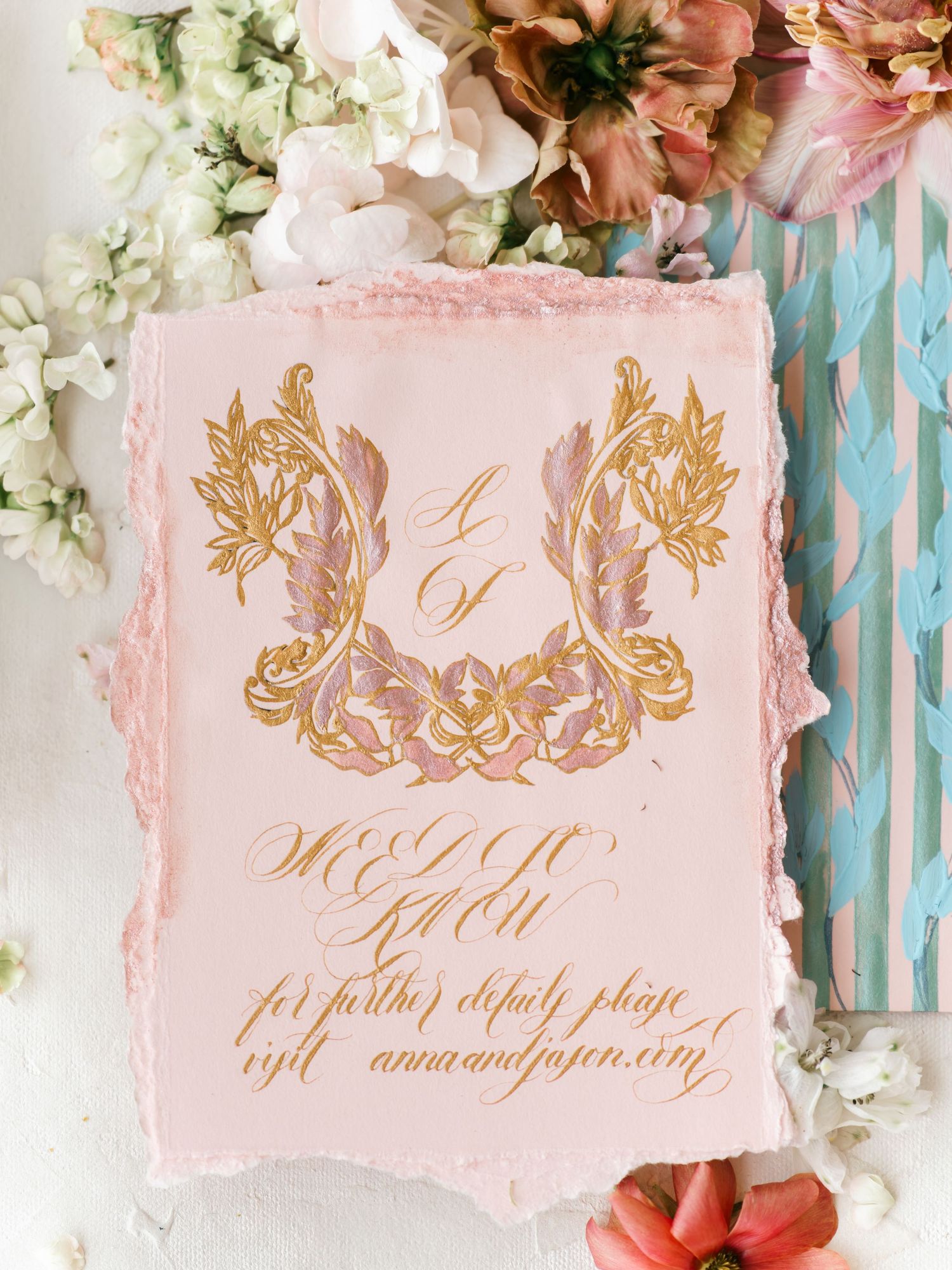 Elegant pink handmade paper wedding invitation with gold calligraphy and ornate floral crest design, surrounded by fresh flowers.