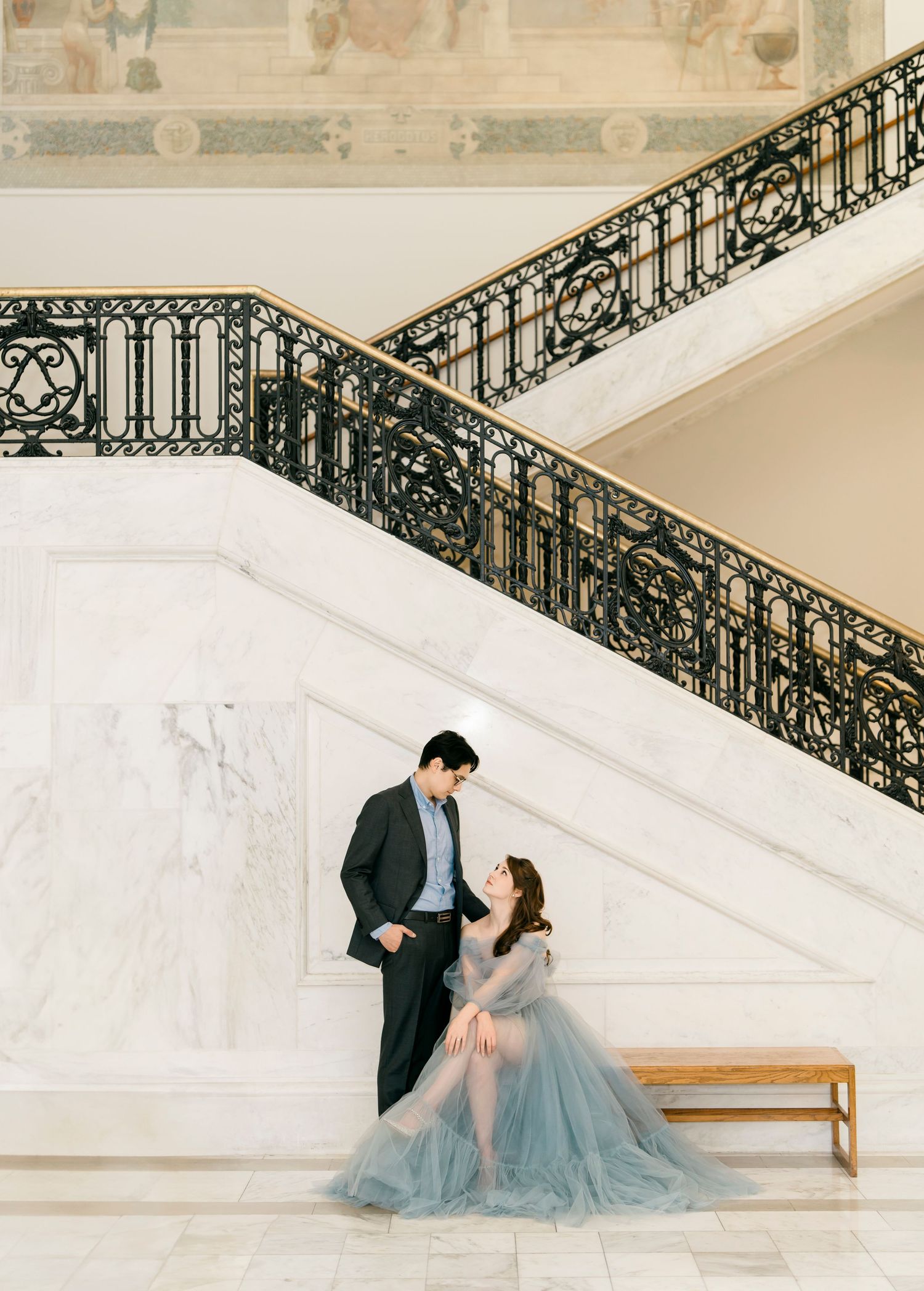 A couple in formal attire poses elegantly on a grand white staircase with ornate black iron railings.