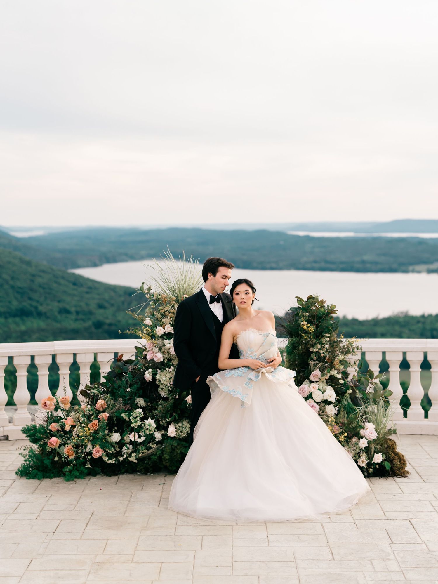 Bride and groom pose on a stone terrace with lush floral arrangements and a scenic lake and hillside backdrop.