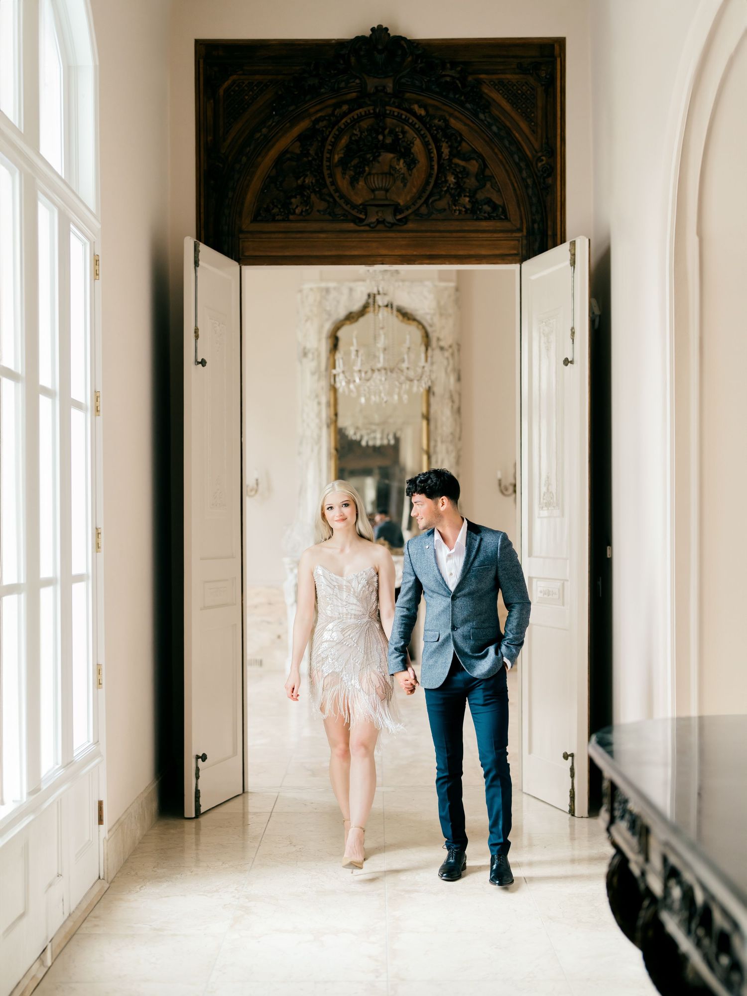 Elegant couple walks through ornate white hallway with tall windows and decorative moldings in wedding attire.