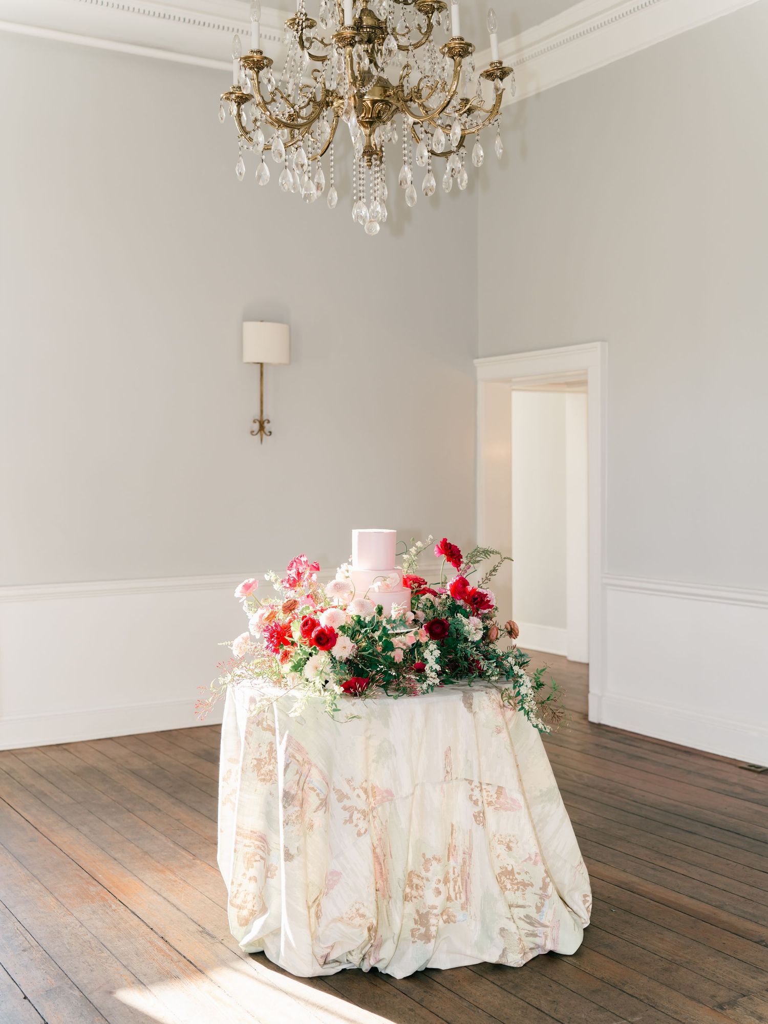 Elegant empty room with crystal chandeliers, white walls, hardwood floors and draped round tables with floral centerpieces.