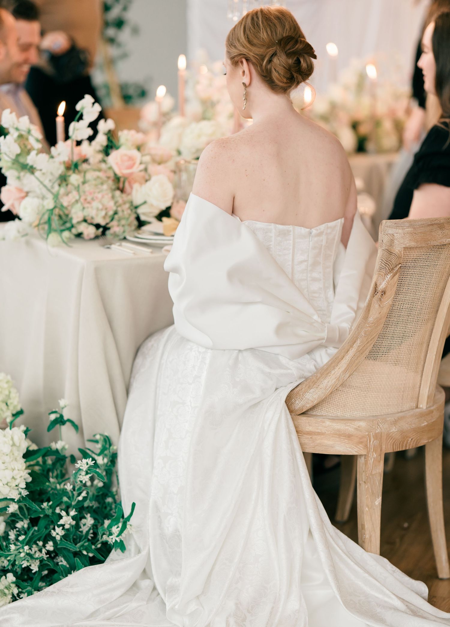 A bride in a flowing white gown sits elegantly in a chair surrounded by romantic floral decorations.