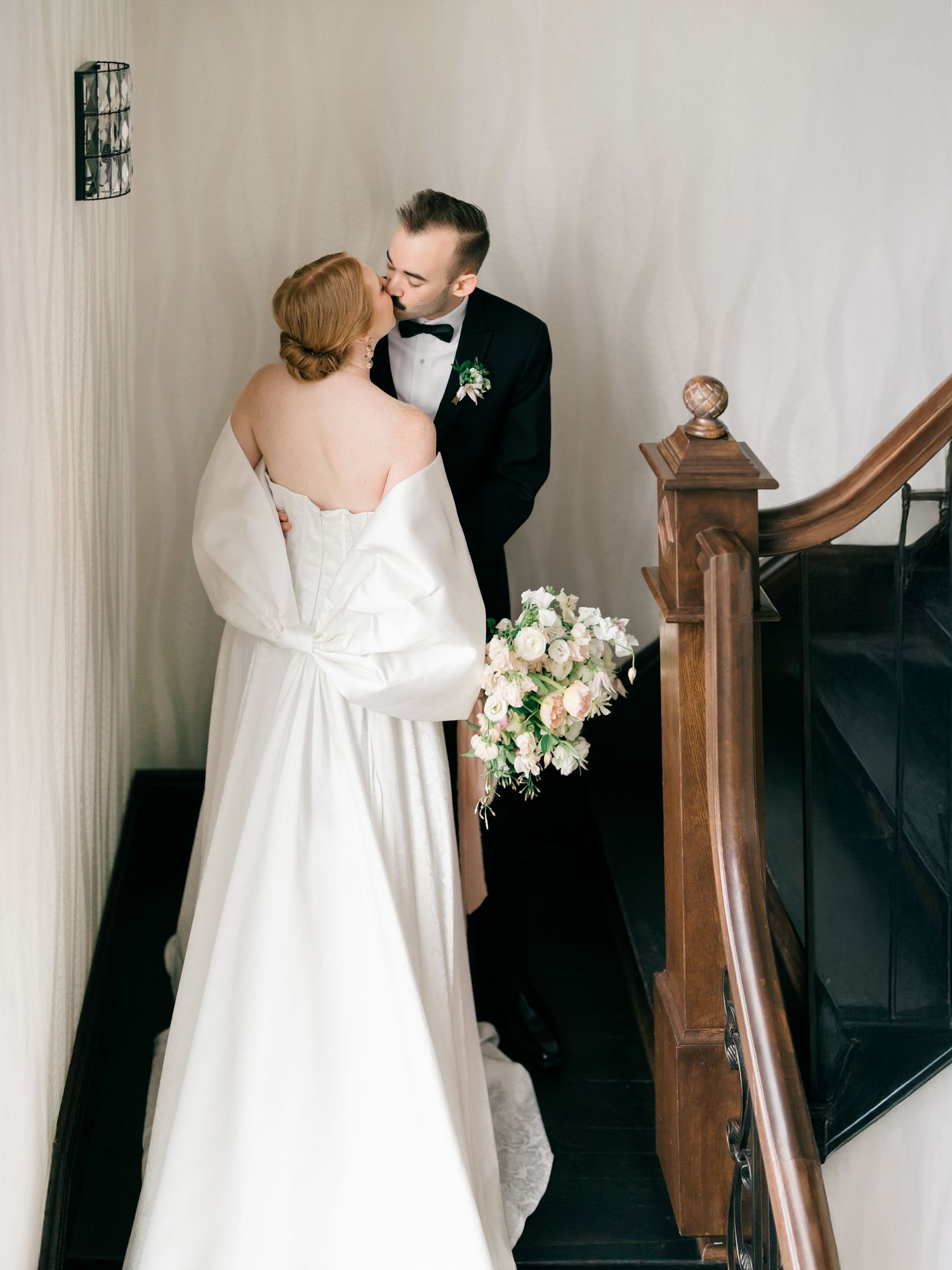 Couple in wedding attire share an intimate moment on a staircase with antique wooden railings and a white floral bouquet.