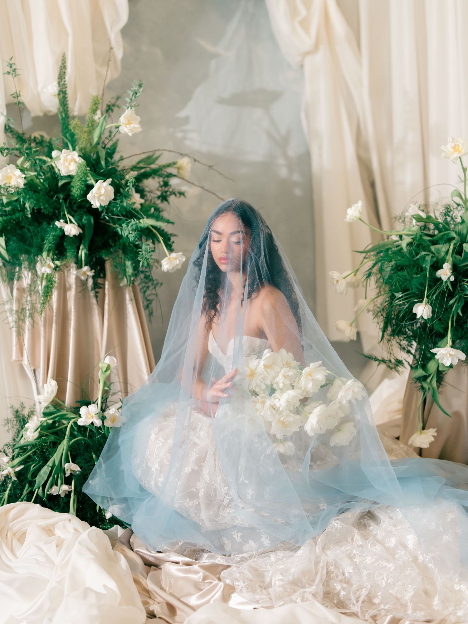 Bride in a blue tulle gown with veil, holding white bouquet, surrounded by lush green floral arrangements.