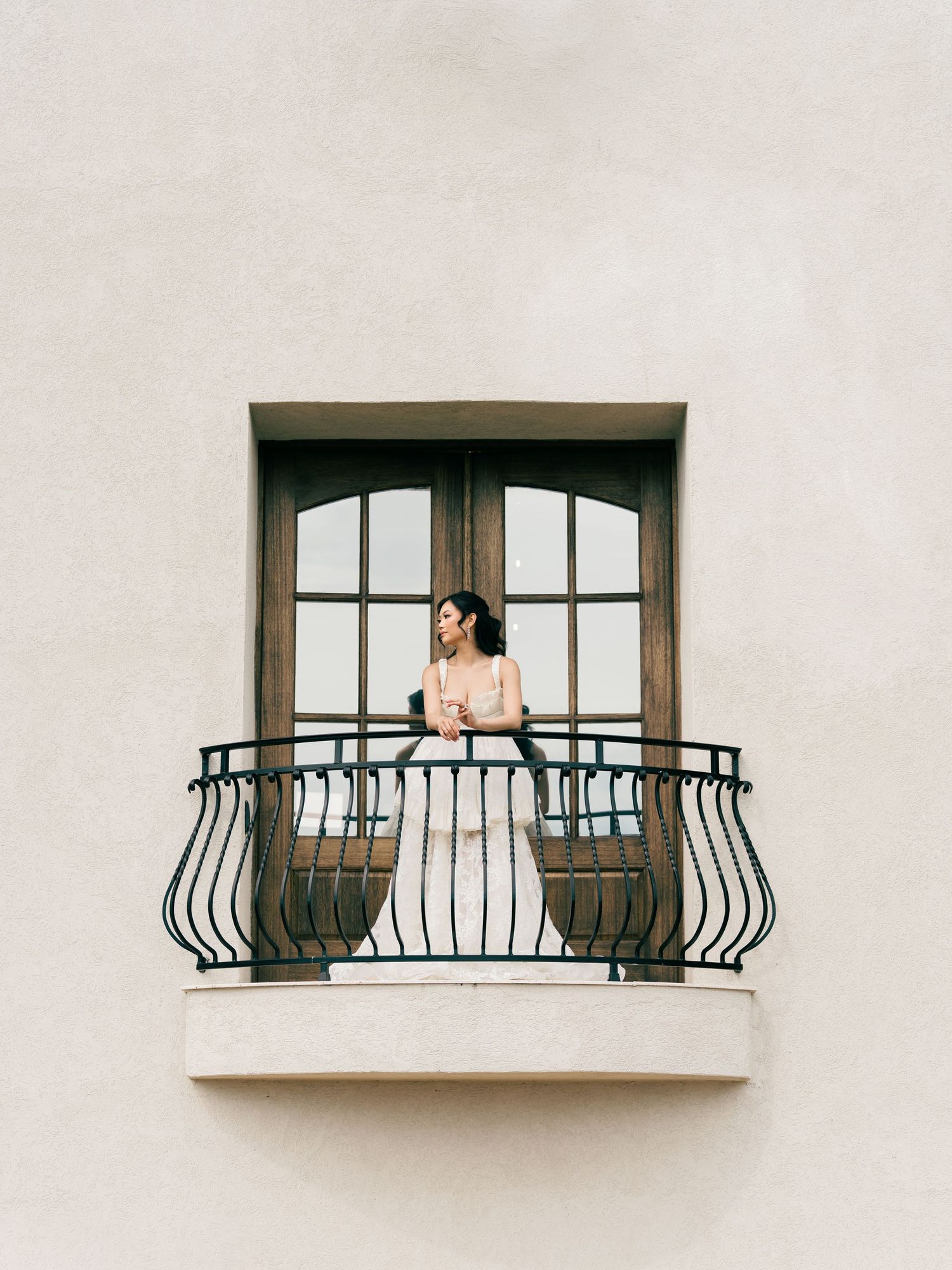 Bride in white wedding dress standing on ornate iron balcony of a minimalist beige building.
