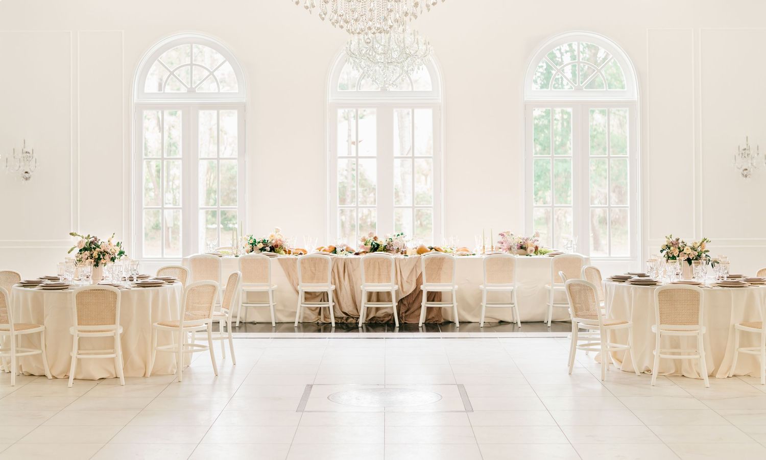 Elegant white wedding reception venue with arched windows, cream-colored chairs, and round tables set for dining.