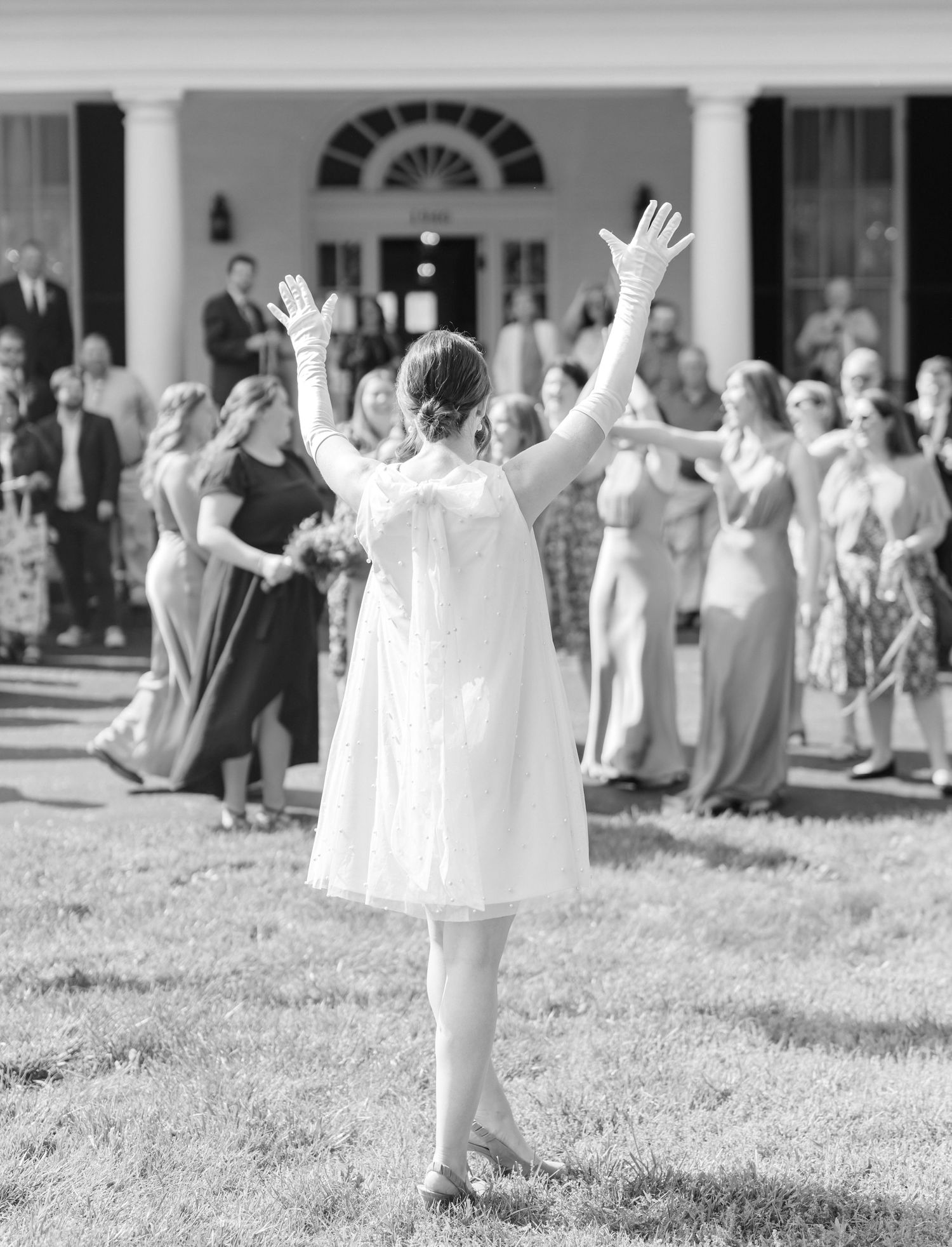 Woman in white dress raises her arms in celebration on a lawn with guests gathered outside an elegant building.