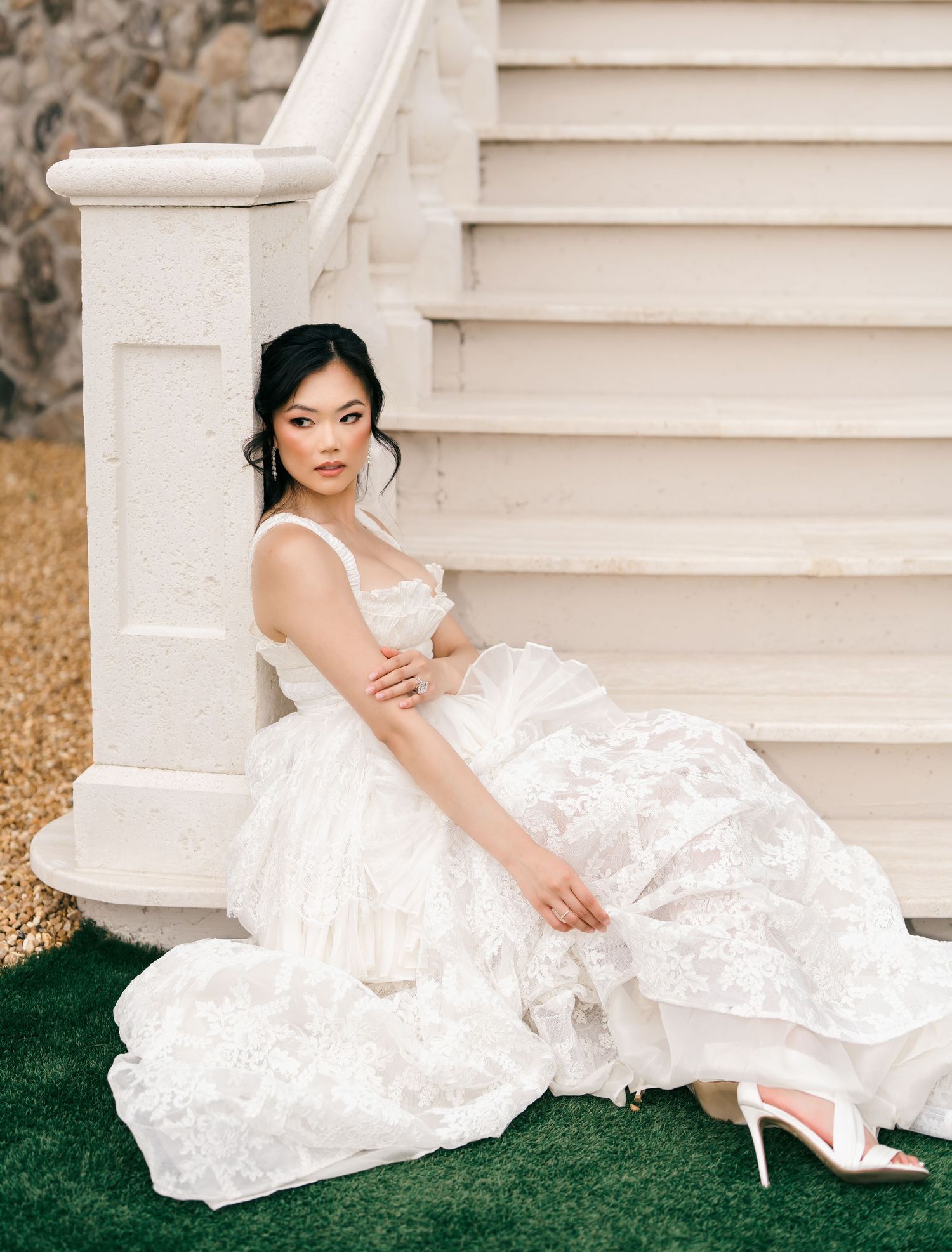 Beautiful bride in layered lace wedding gown seated on white staircase steps with strappy heels.