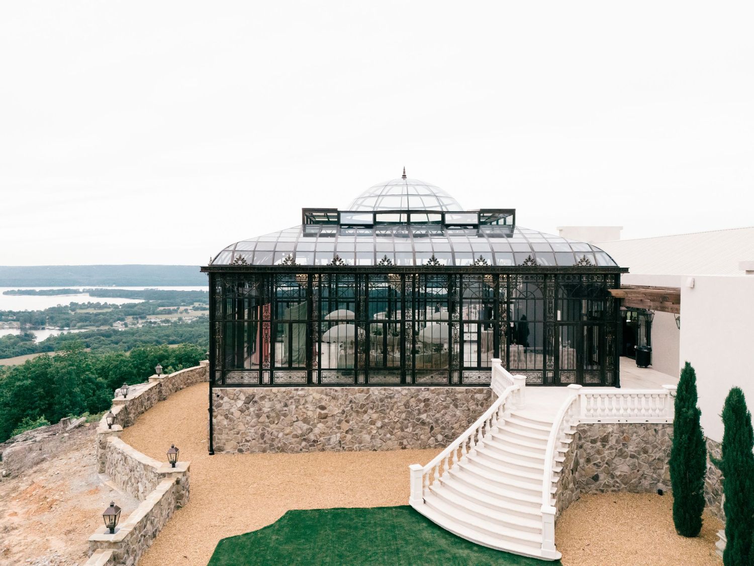 Elegant glass greenhouse conservatory on a hilltop with white staircases and panoramic water views.