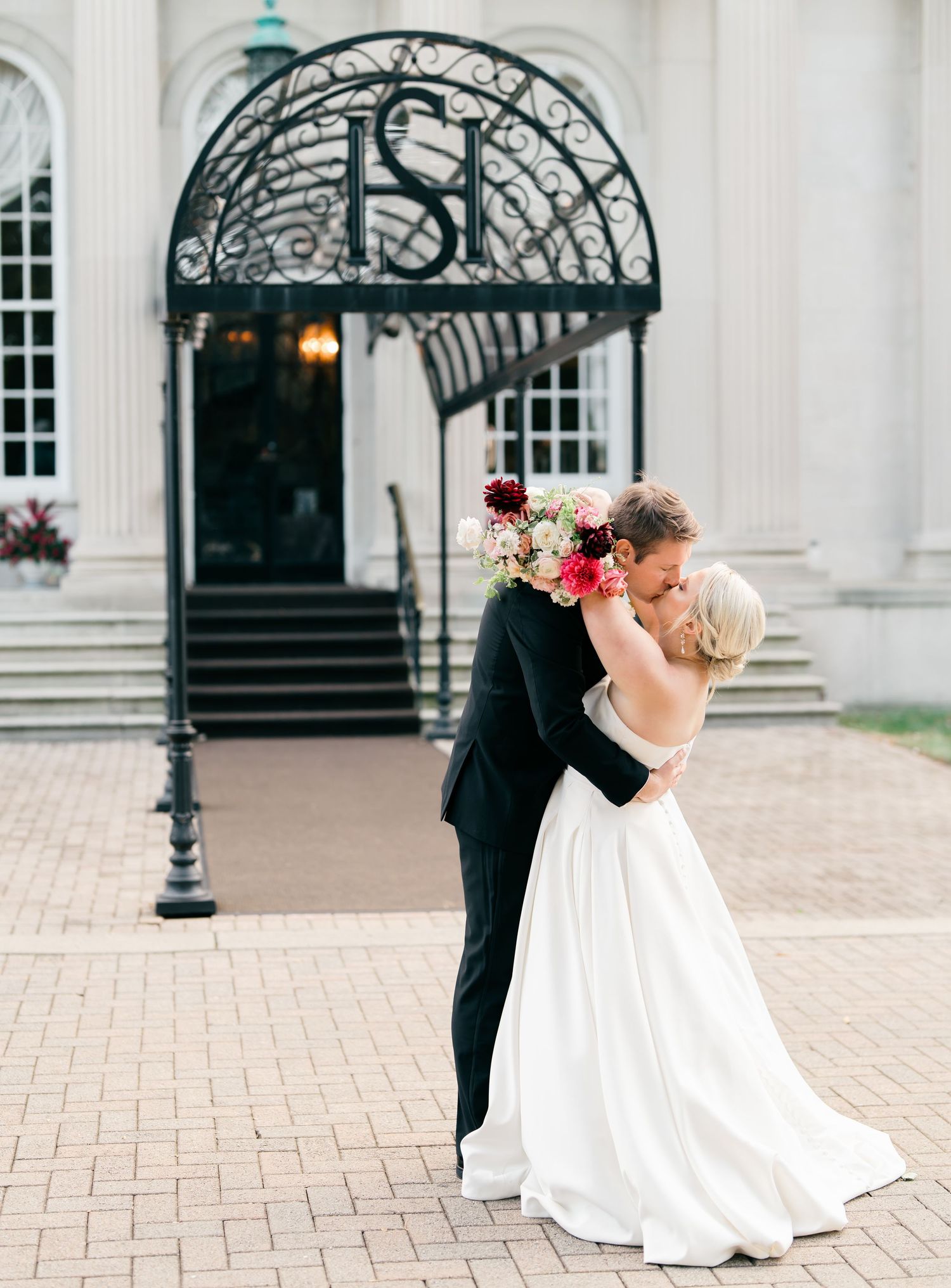 Elegant couple embraces under ornate wrought iron entrance with classical columns and brick path at historic venue.