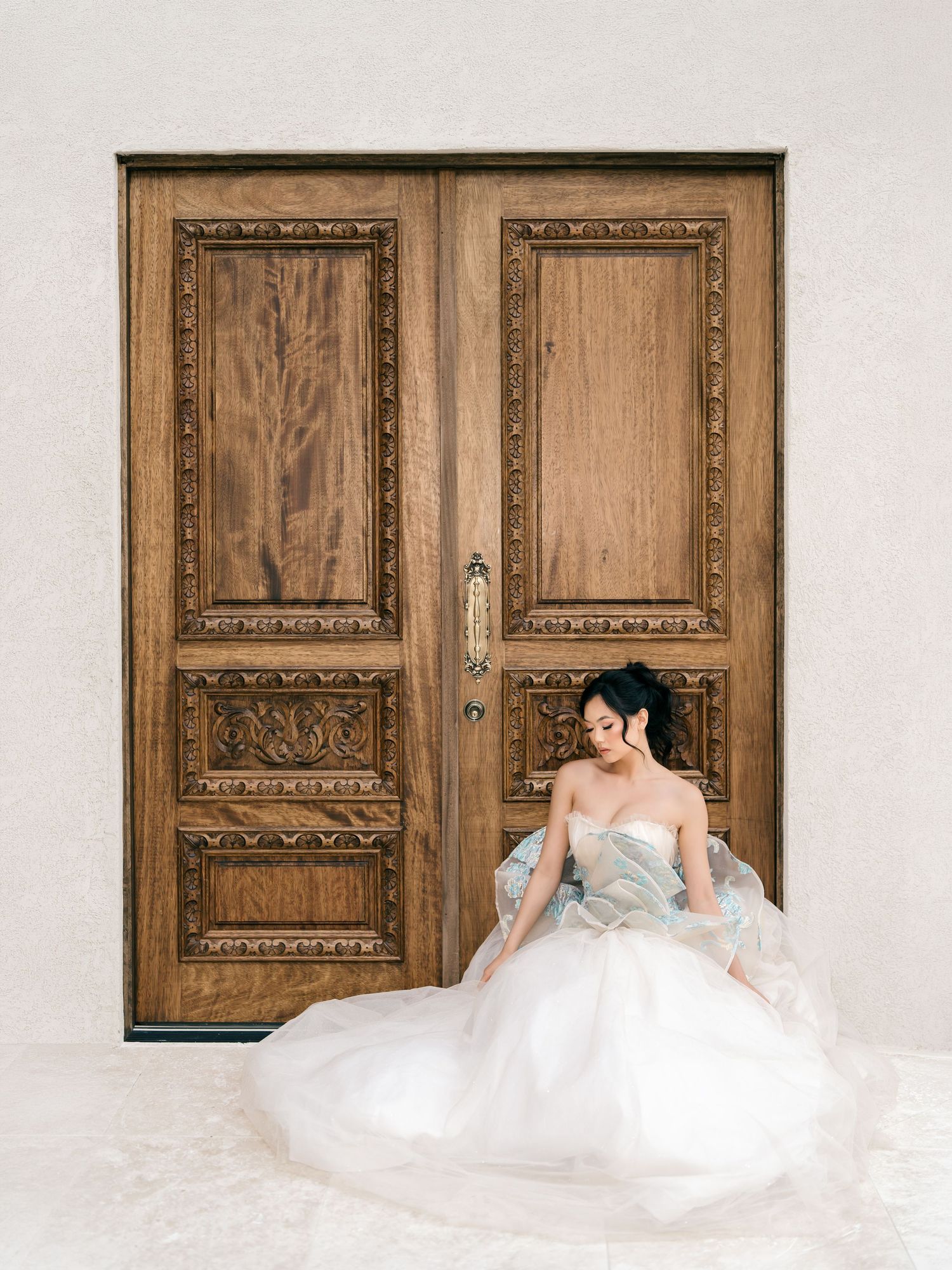 Bride in strapless white ballgown sitting gracefully before ornate carved wooden double doors.