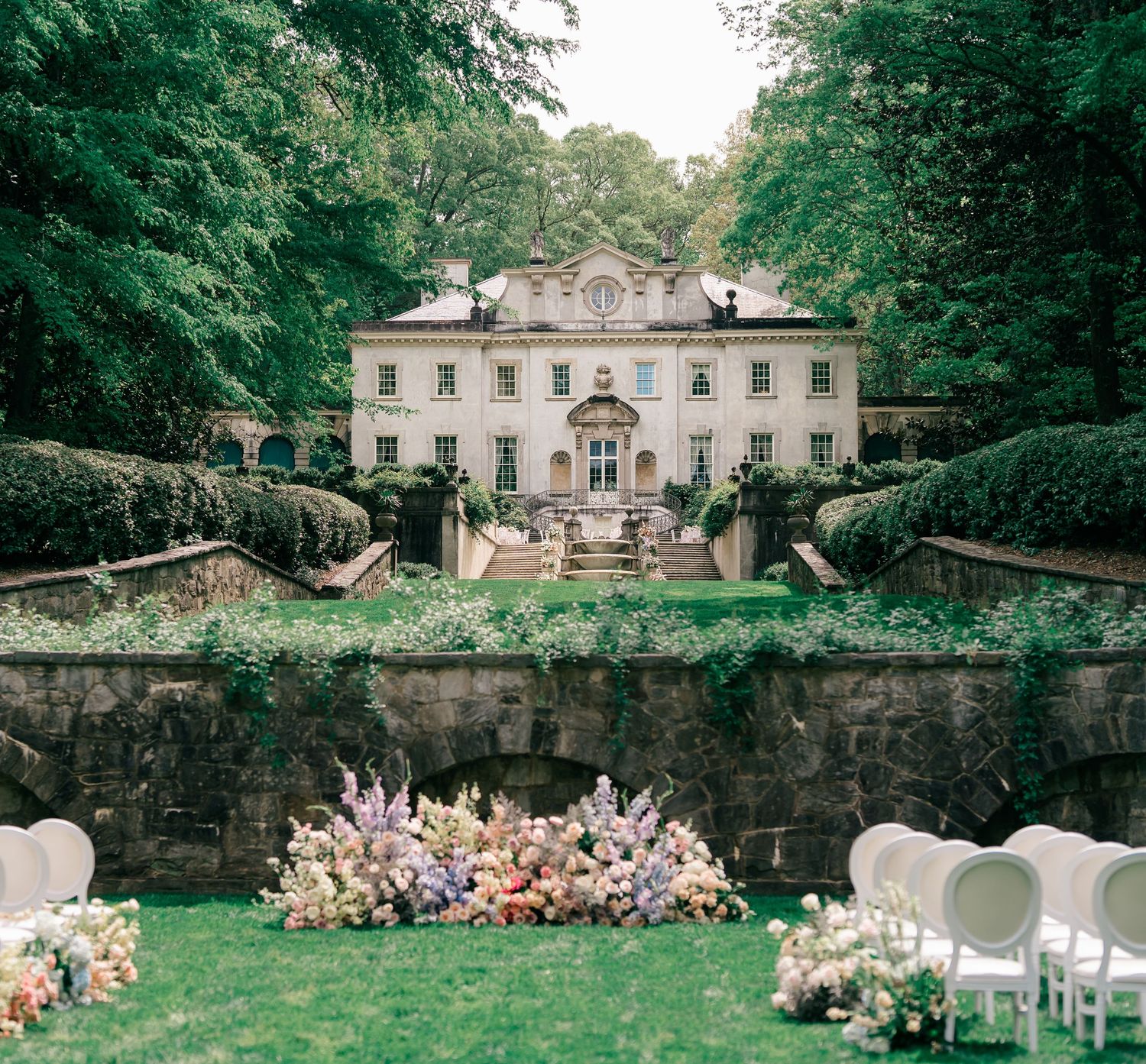 Elegant outdoor wedding ceremony at a grand white manor house with lush gardens and colorful floral arrangements.