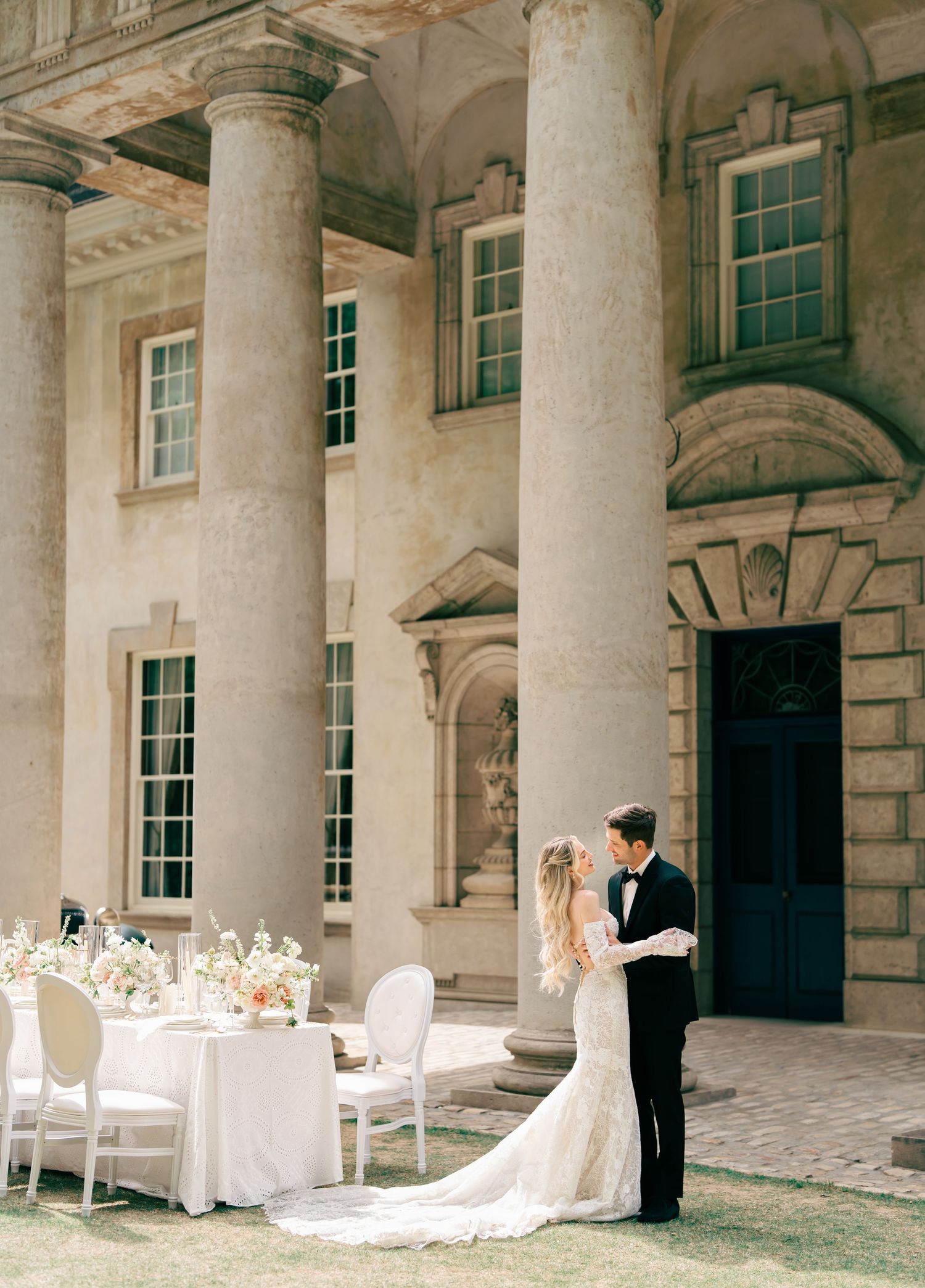 Bride and groom embrace outside elegant neoclassical venue with grand columns and outdoor reception table.
