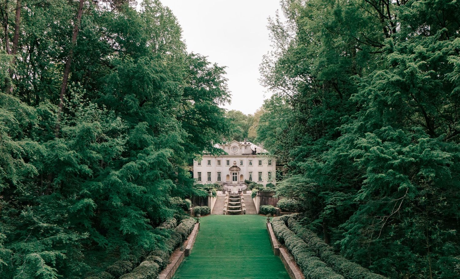 Elegant white mansion surrounded by lush green trees with a manicured lawn pathway leading to grand entrance steps.